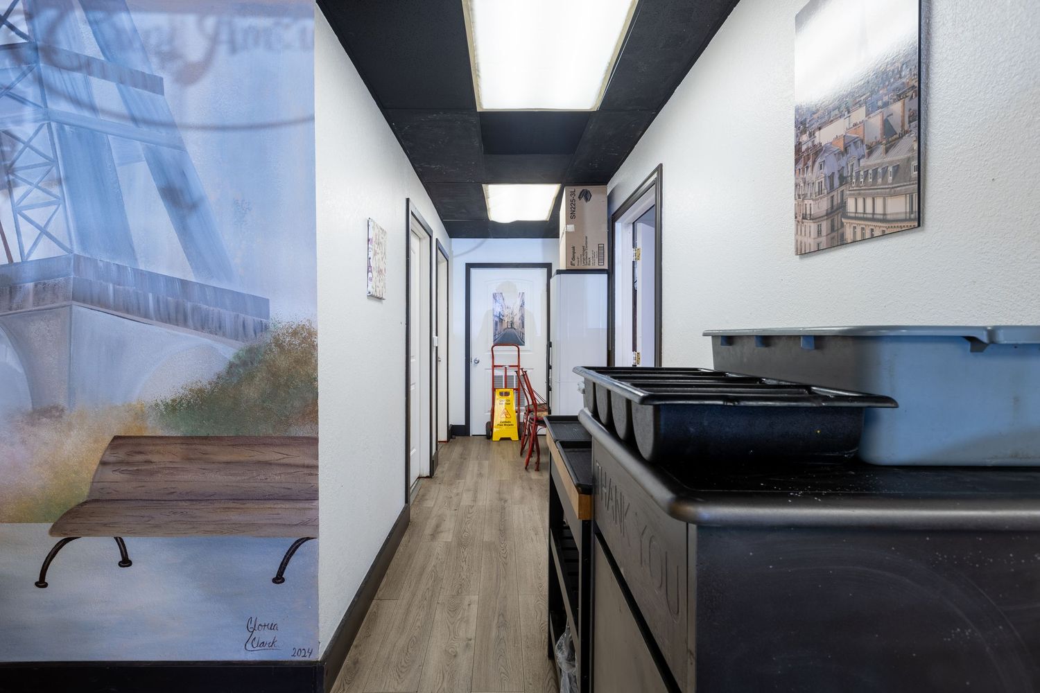 Narrow commercial hallway with hardwood floors, artwork on walls, black countertop, and a yellow caution sign in the background.