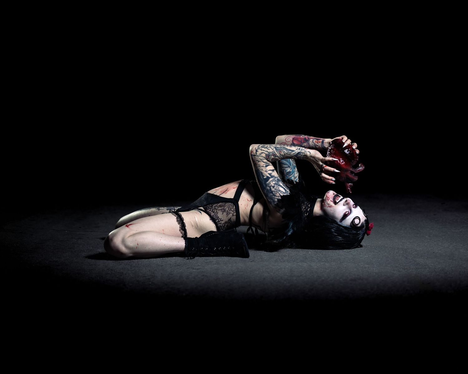 Tattooed woman in black lingerie lying dramatically on dark floor in artistic gothic pose.