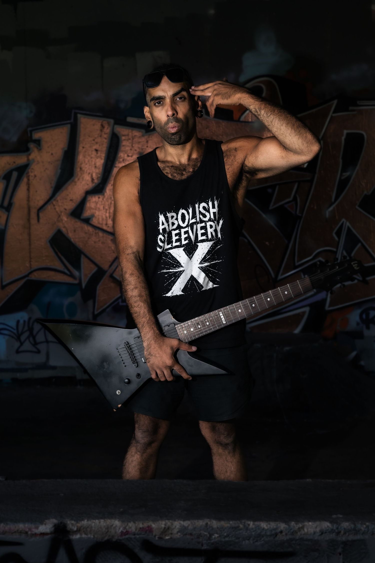 Musician in dark lighting plays electric guitar while wearing a black t-shirt with text against an industrial backdrop.