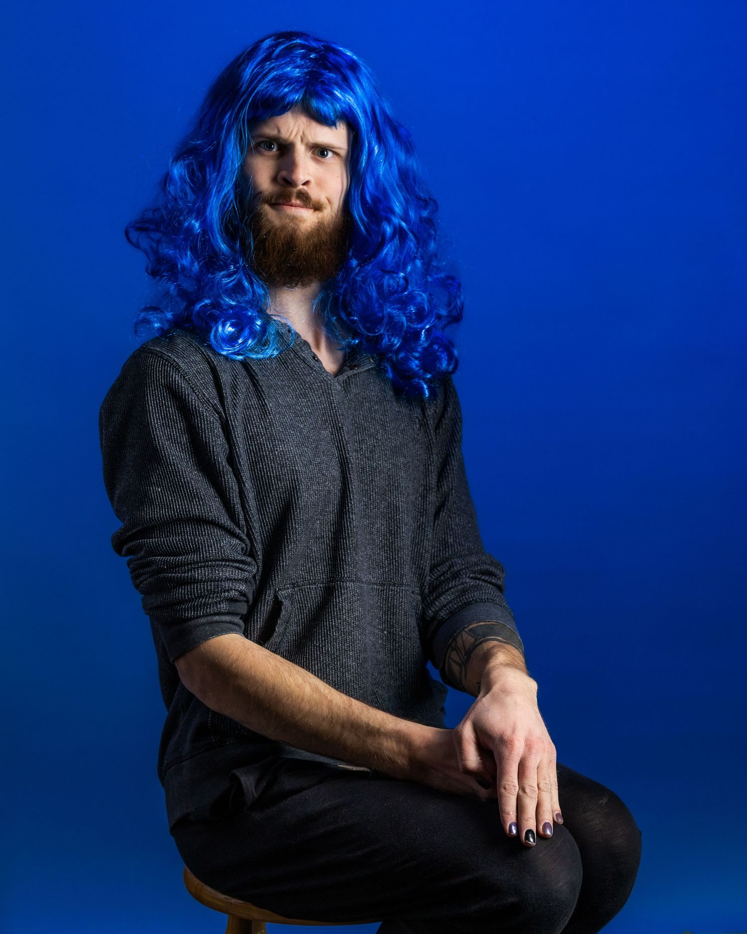Person wearing a bright blue wig and gray sweater poses against a vibrant blue background.