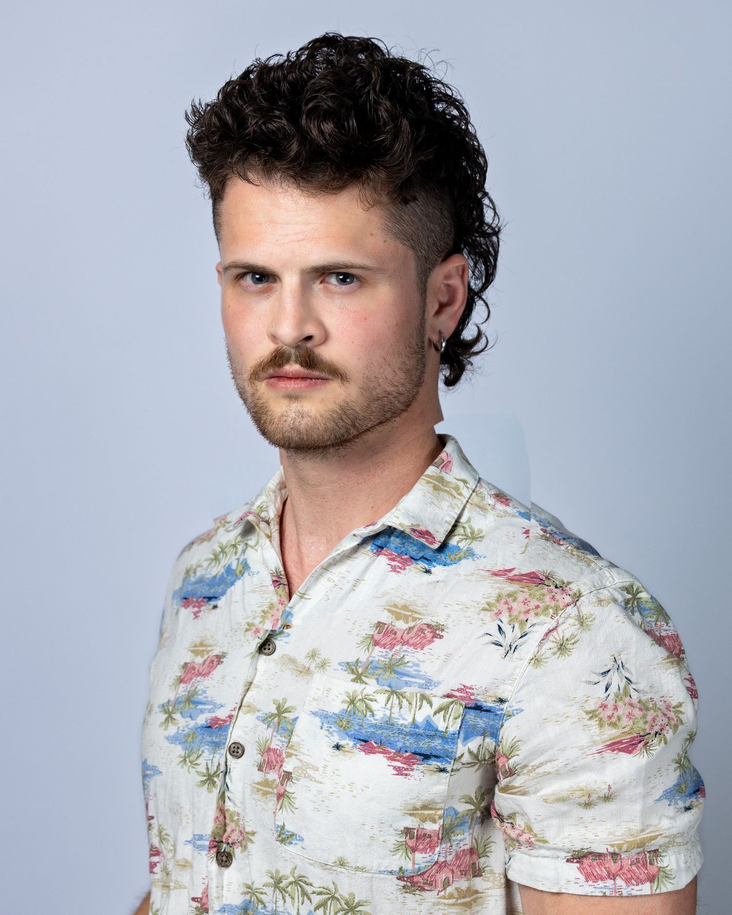 Young man with curly dark hair, mustache, ear gauges, wearing a floral Hawaiian shirt, posing against a light background.