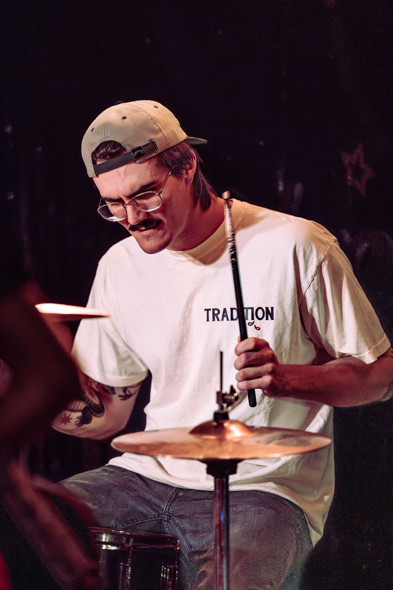 Young drummer with mustache and cap wearing white Tradition t-shirt playing drums on stage.