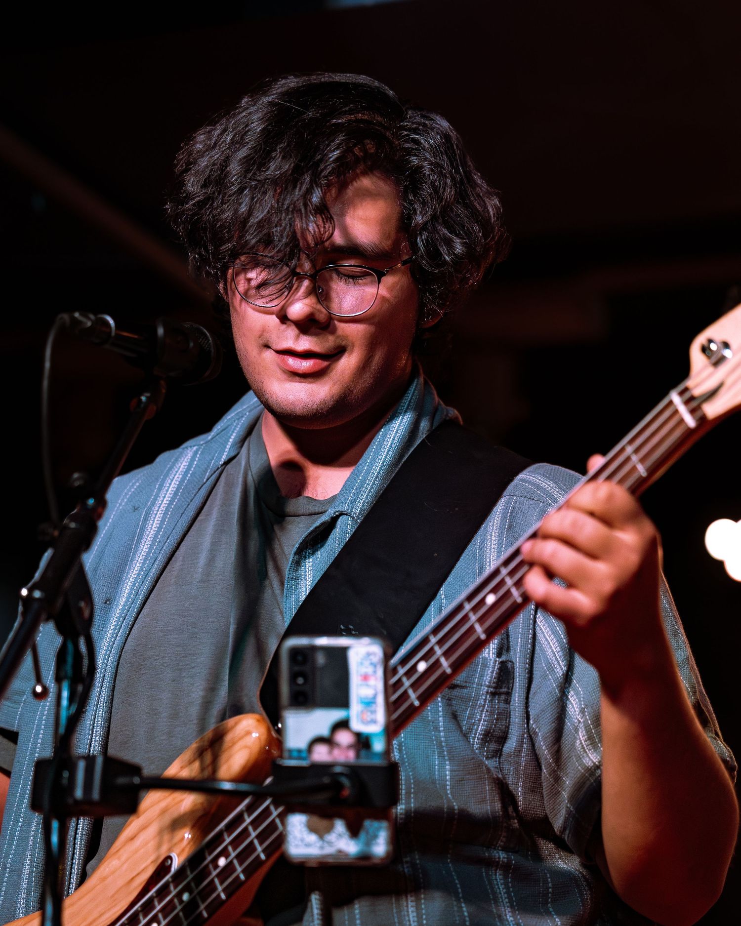 Young musician with glasses playing bass guitar on stage under warm lighting at a live music venue.