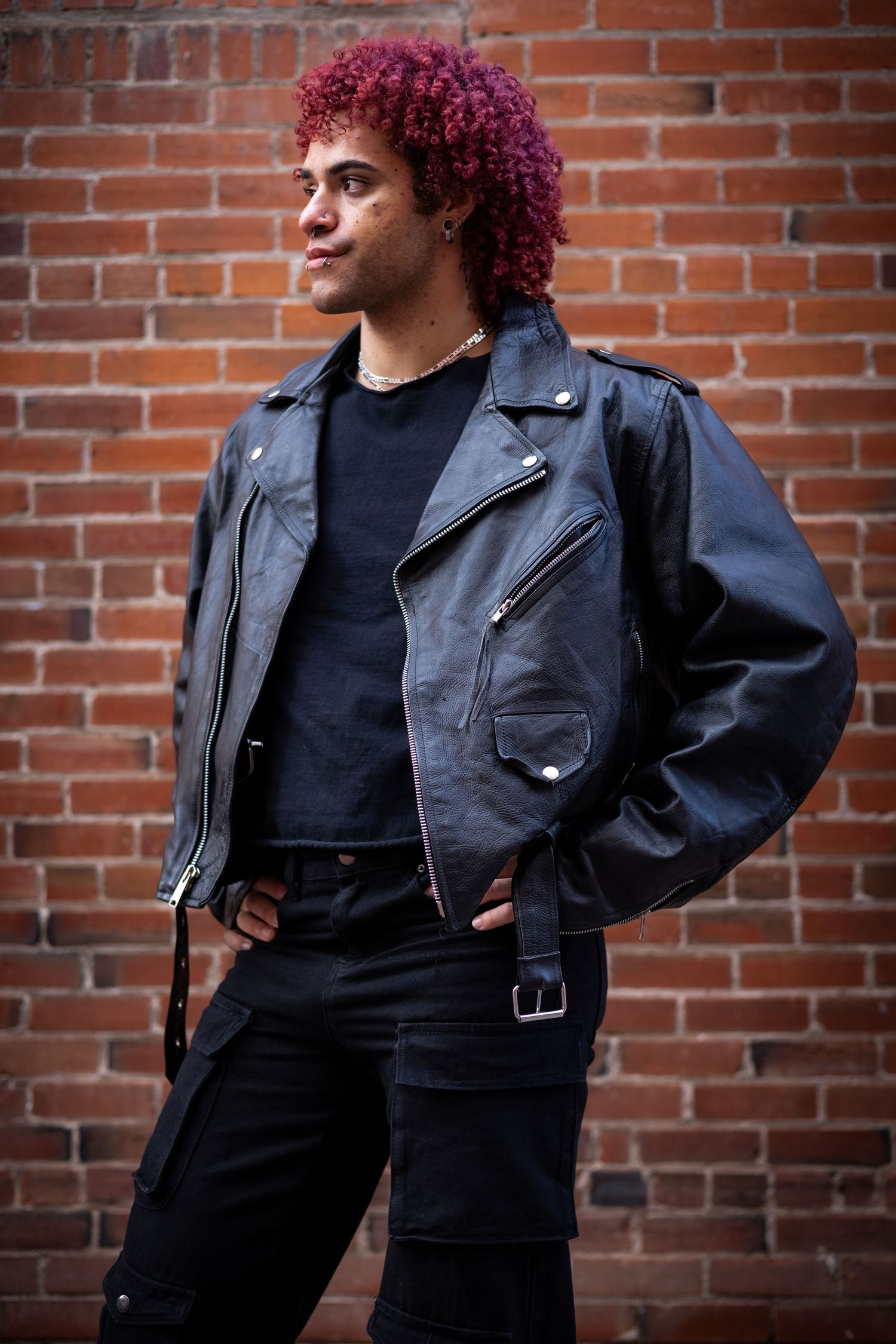 Edgy street style with magenta curly hair, black leather jacket and dark outfit against brick wall.