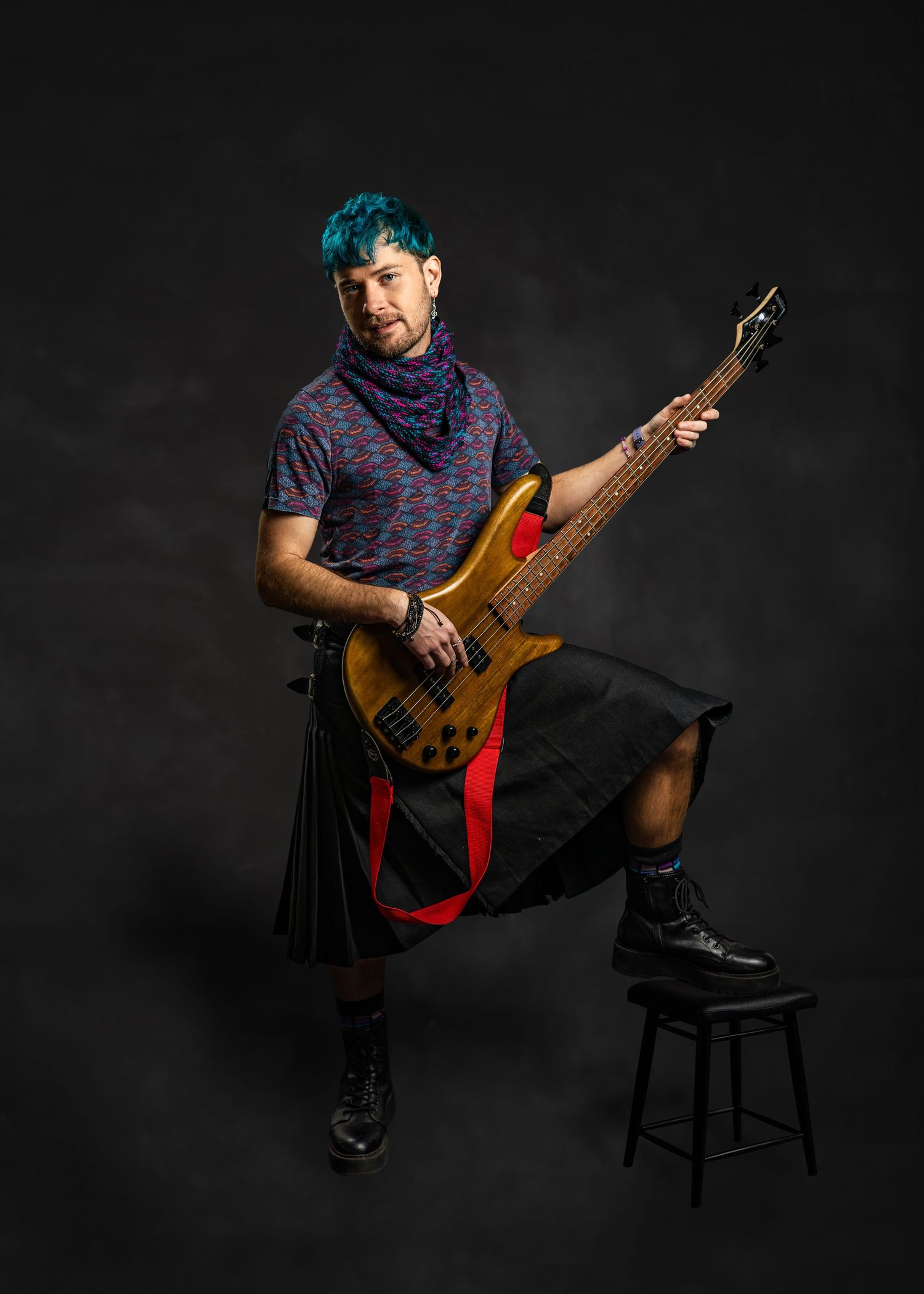 Musician with teal hair plays bass guitar wearing a kilt and blue scarf in a dark studio.