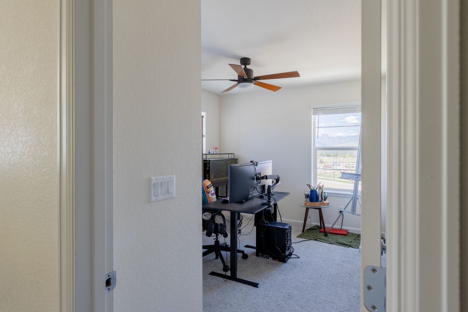 Home office with ceiling fan, desk, chair, and natural light from windows, featuring neutral carpet and white walls.