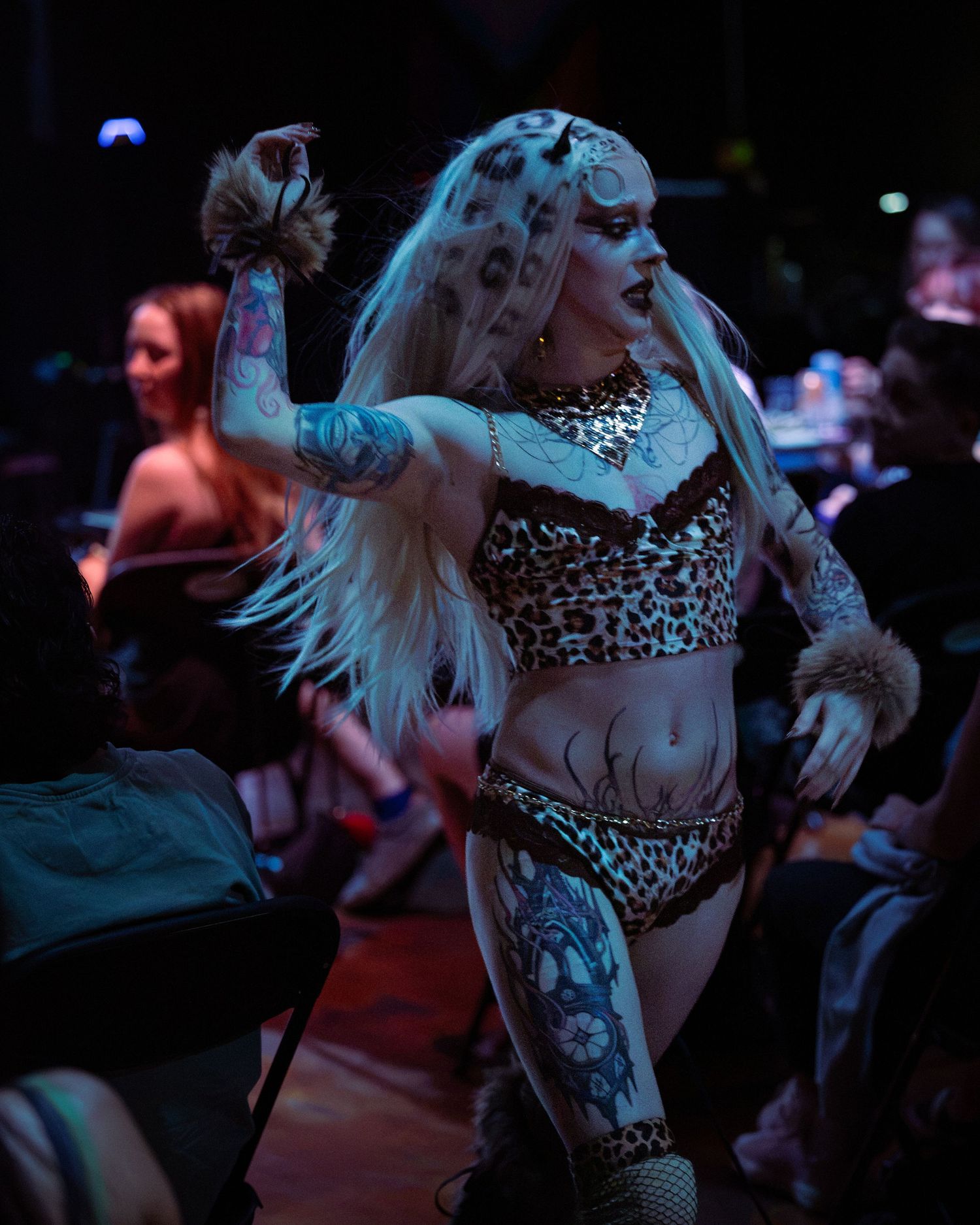 Heavily tattooed performer in leopard print costume dances energetically among audience under colorful stage lighting.