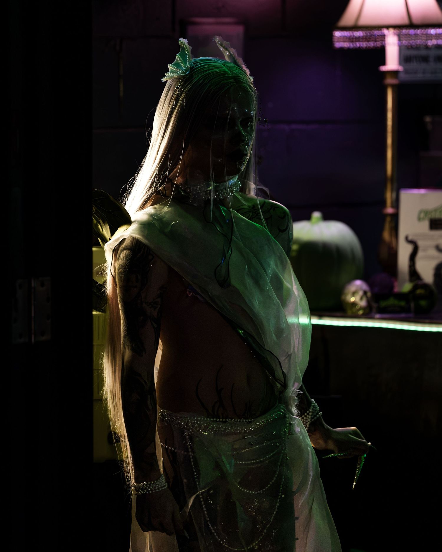 A tattooed woman in a white costume with green feather headpiece poses in dramatic dark lighting.