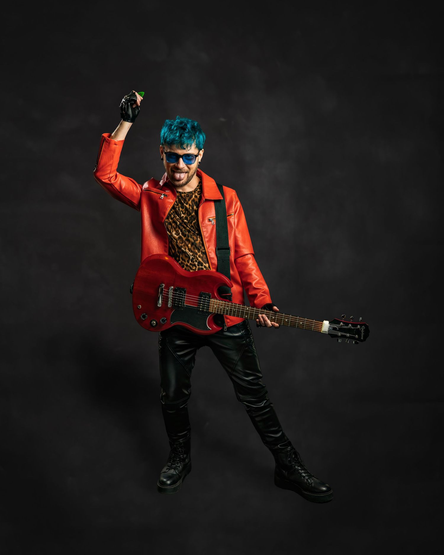 Teal-haired guitarist in red leather jacket and leopard print shirt playing electric guitar energetically.