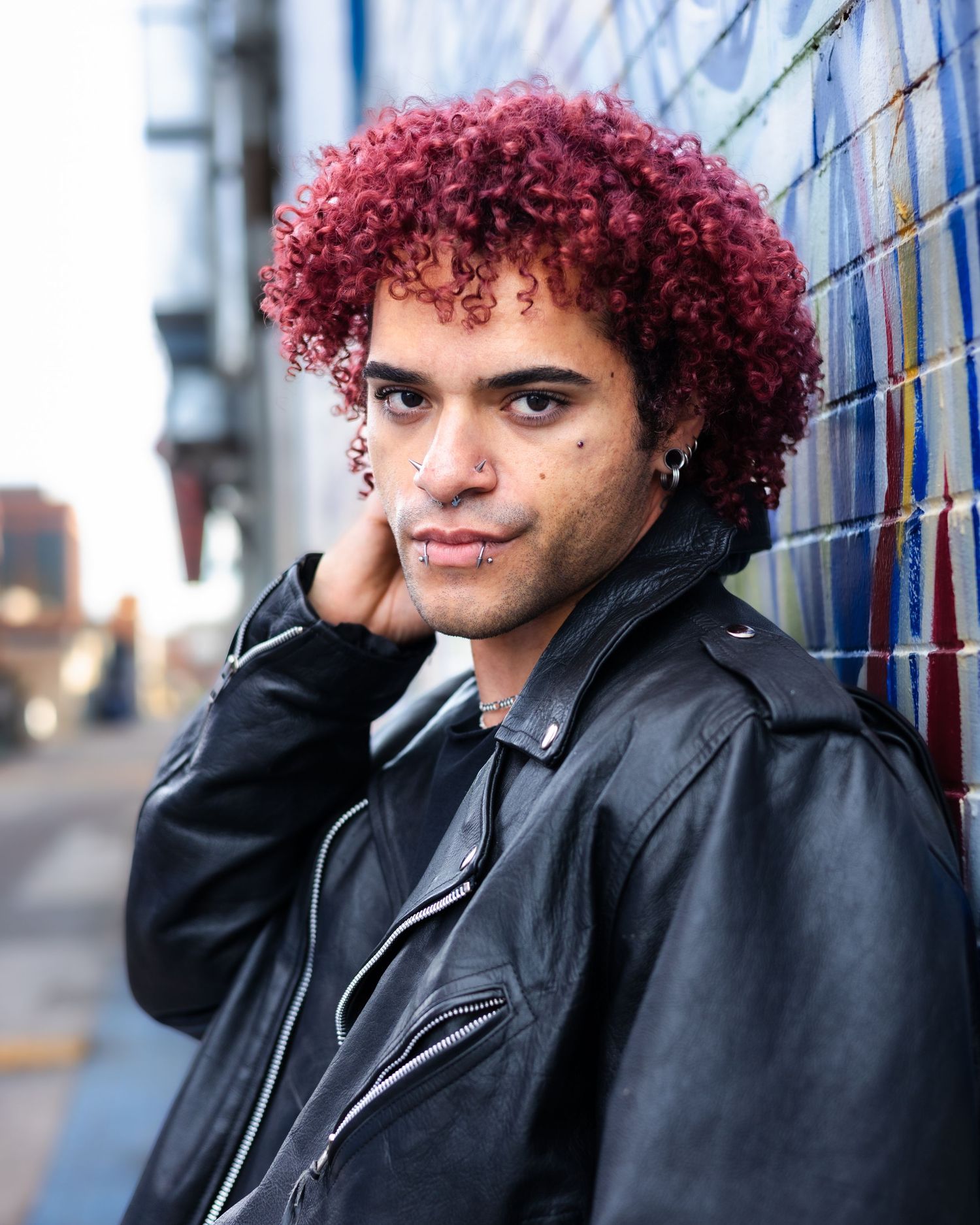 Portrait of person with burgundy curls against urban backdrop.