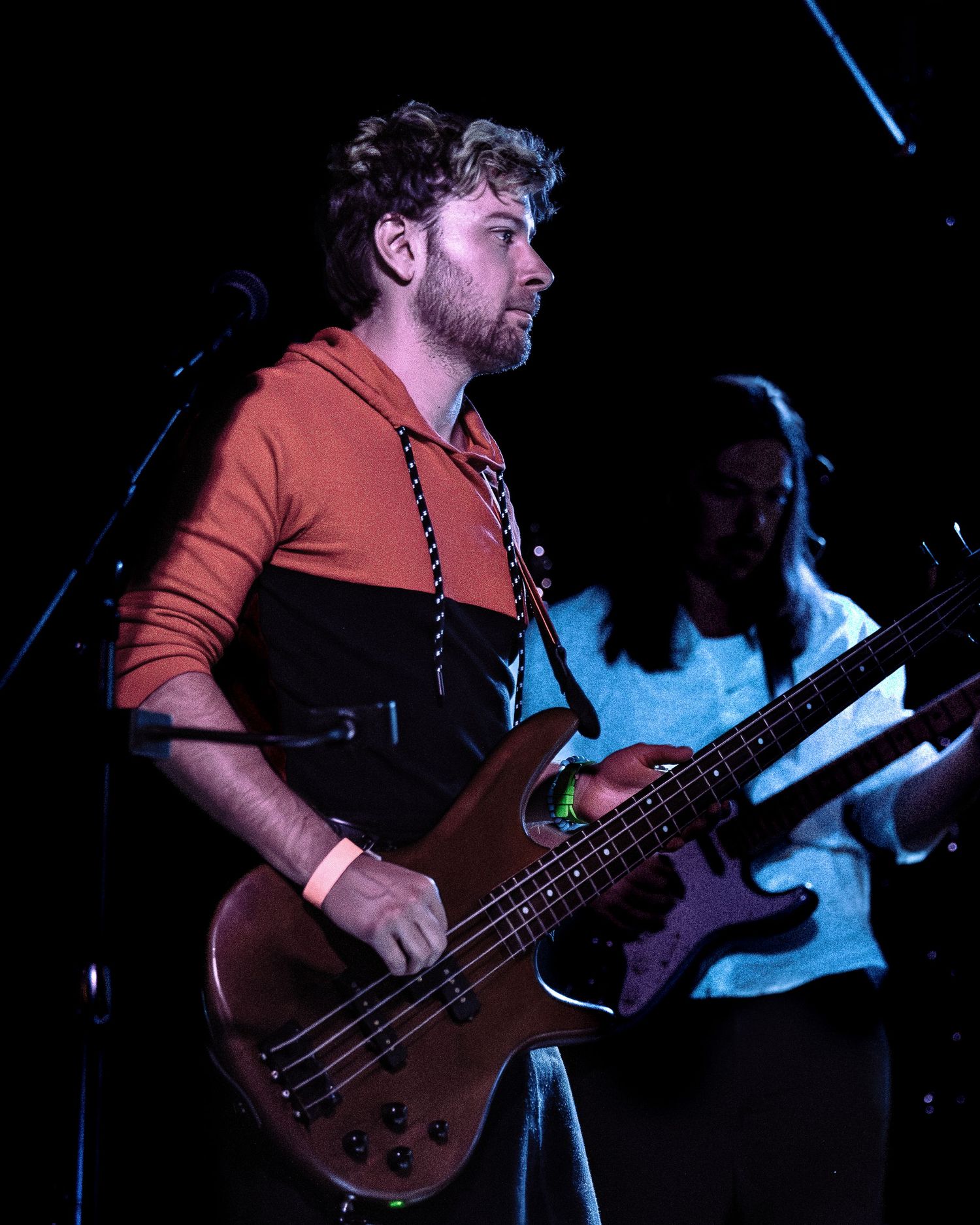 Bearded bass guitarist in orange hoodie performing live on dark stage.
