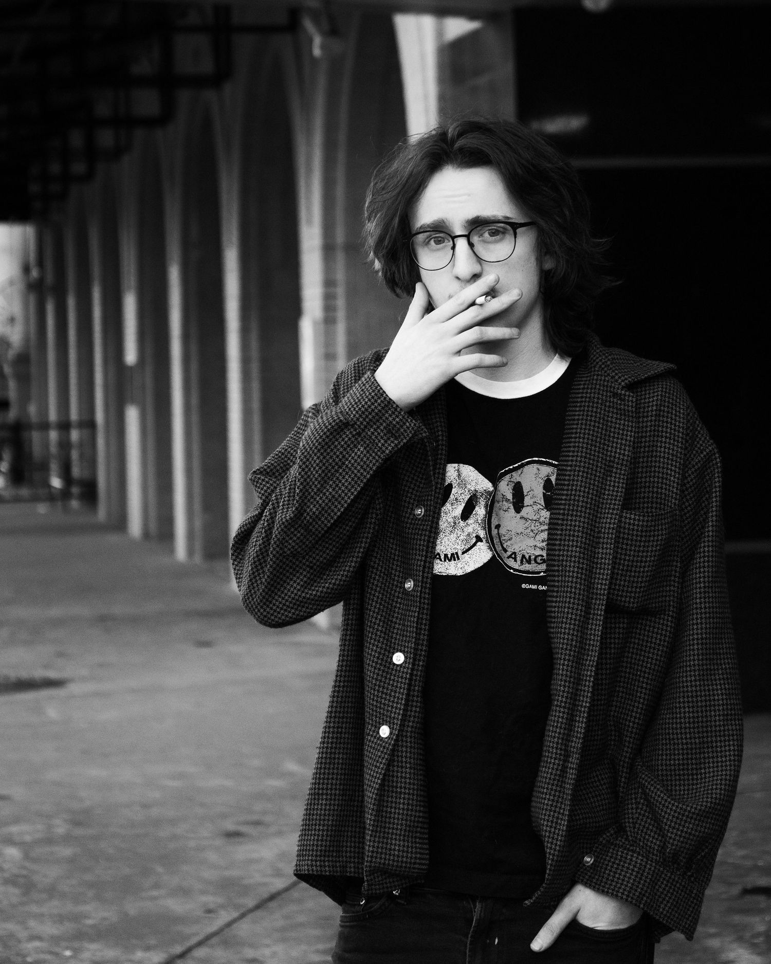 Person in glasses and dark jacket smoking outside in black and white urban setting.