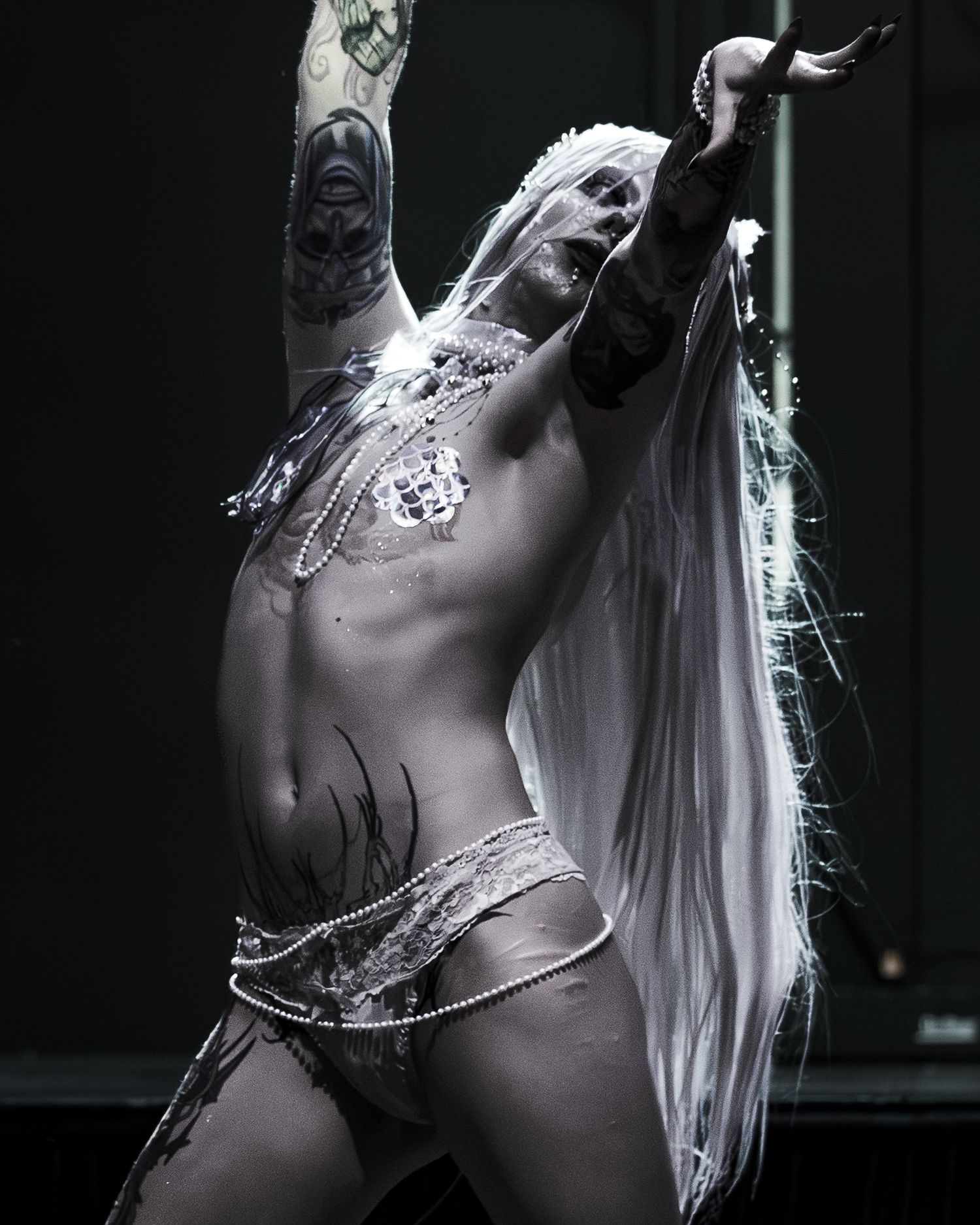 Tattooed performer in silver costume with long white hair raises arms dramatically on dark stage.