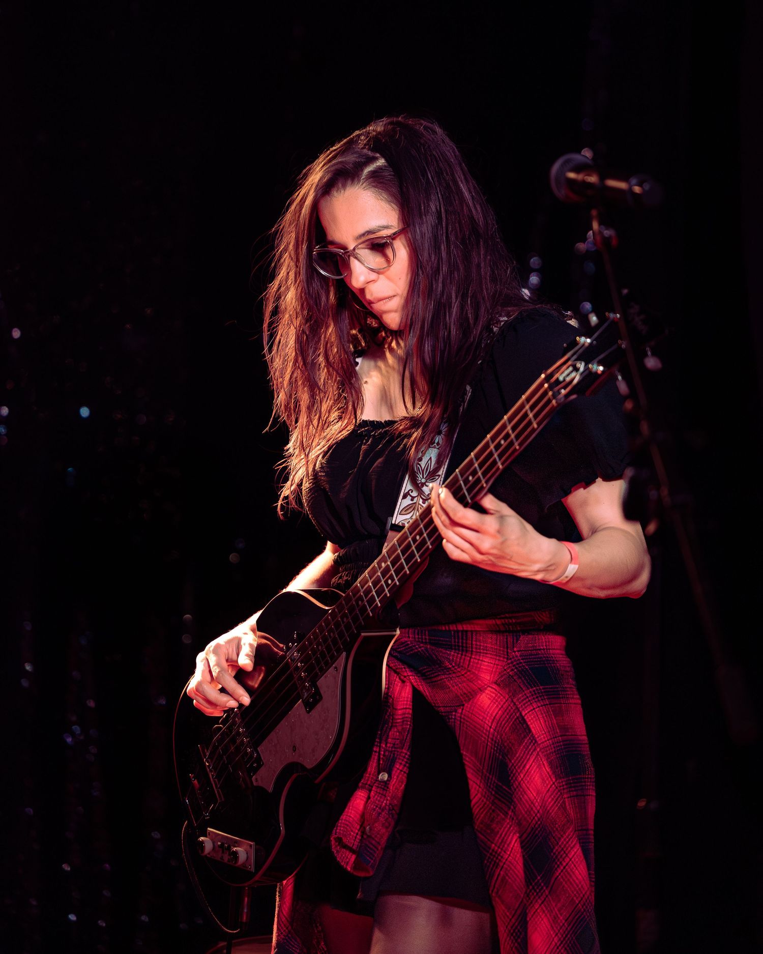 Female guitarist with long dark hair and glasses playing black bass guitar under red stage lighting.