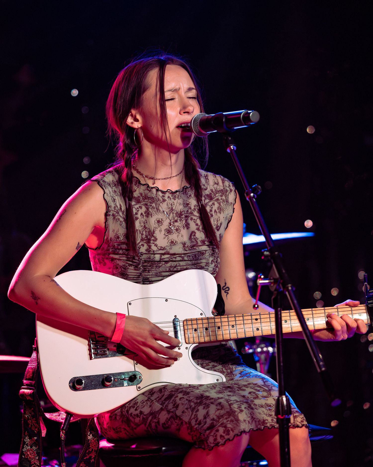 Young woman playing white Telecaster guitar and singing into microphone on stage under pink stage lighting.