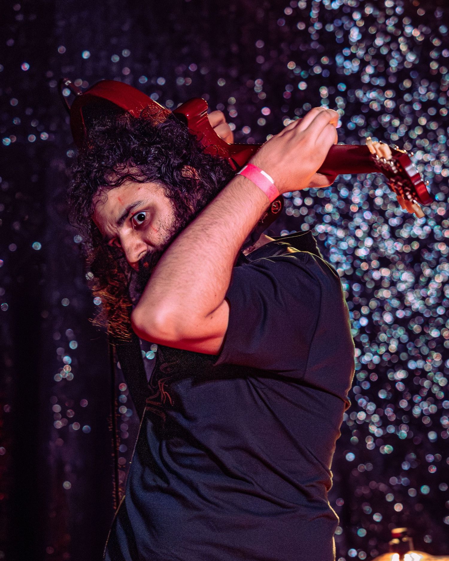 Curly-haired guitarist dramatically raises red electric guitar overhead on a glittering stage.