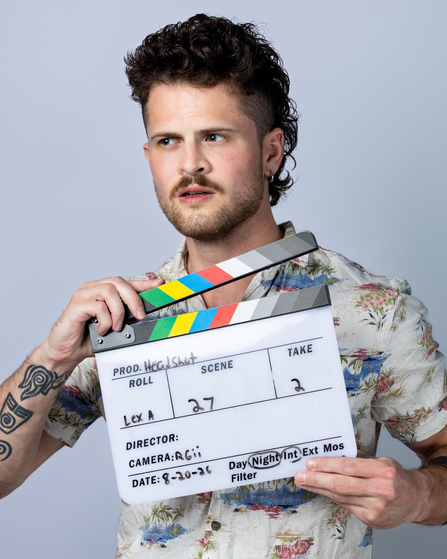 Young man with tattoos holding a film clapperboard on a movie set, wearing a floral shirt.