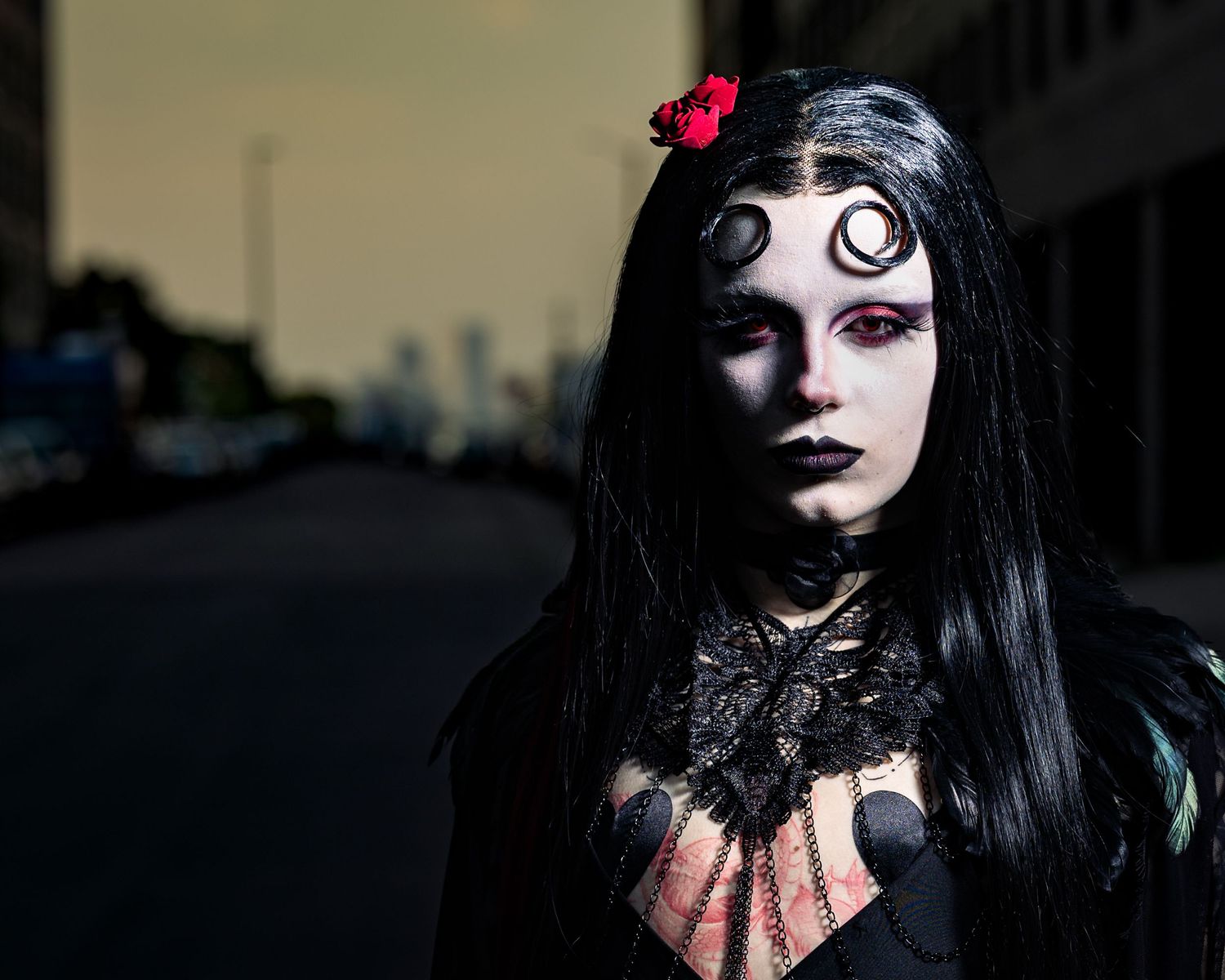 Gothic woman with dramatic makeup, black hair, red flower, and dark lace outfit poses on a city street.