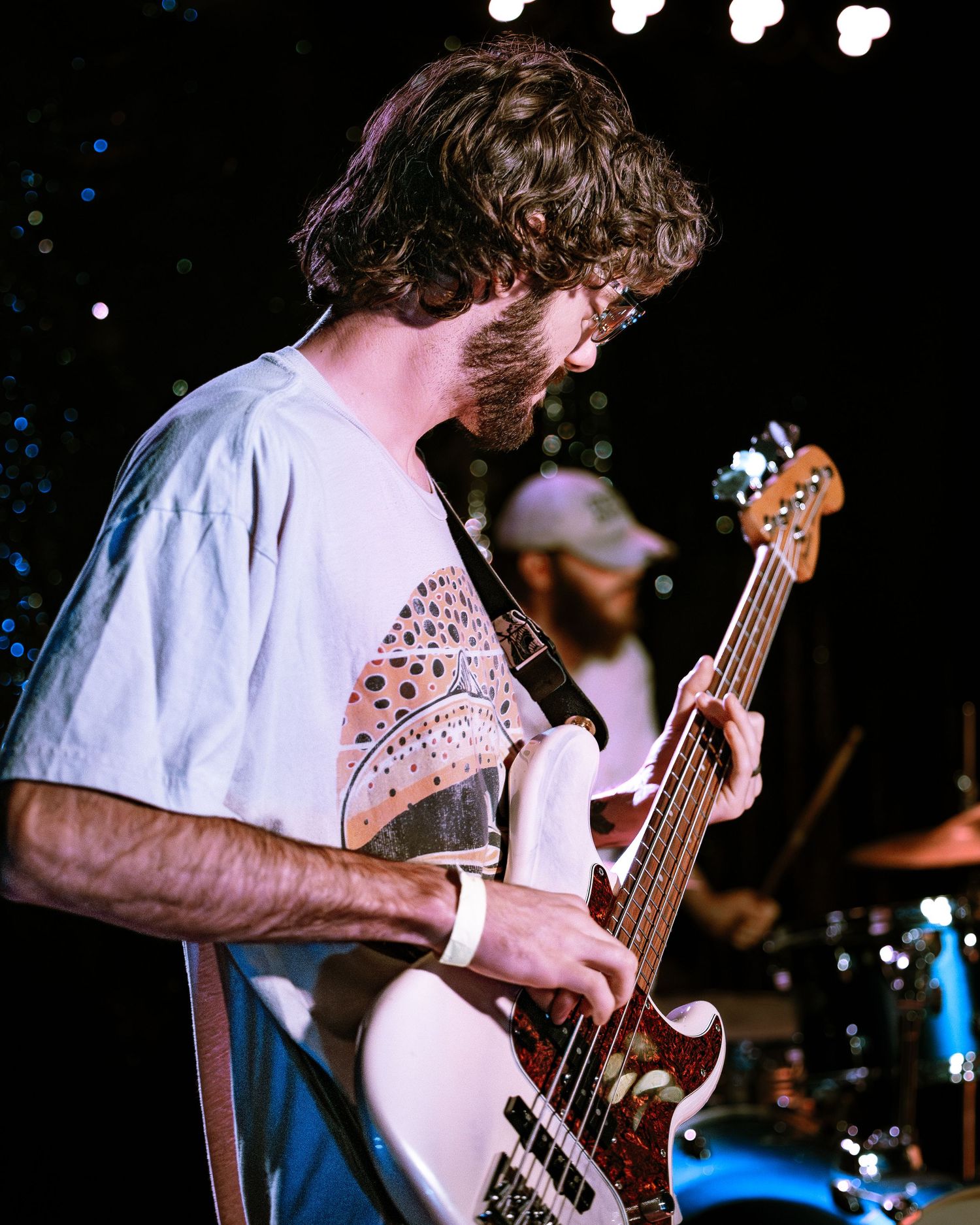 Bearded bassist in white t-shirt plays white bass guitar on dark stage.