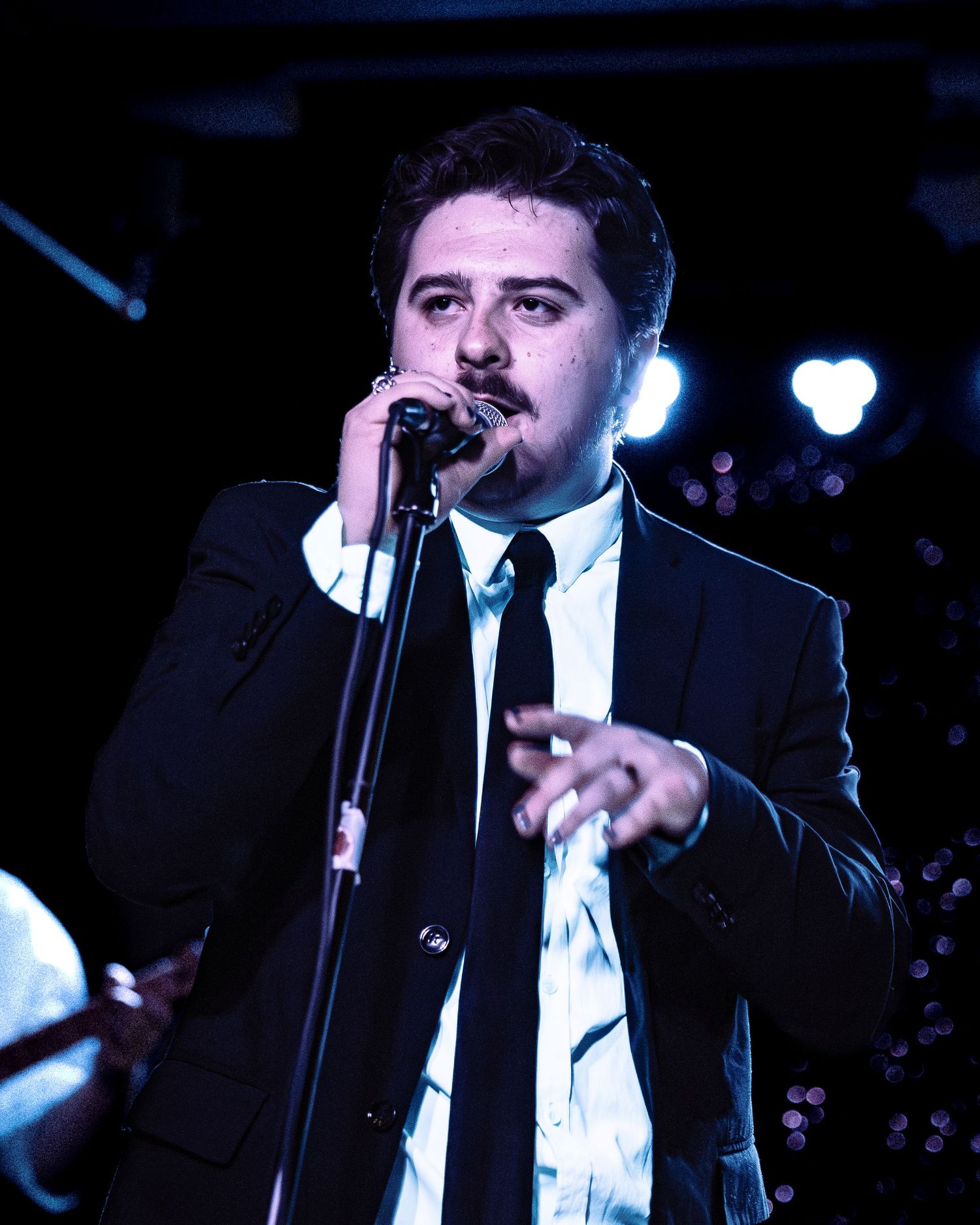 A male singer in a black suit and tie performs on stage, holding a microphone at a dimly lit venue.