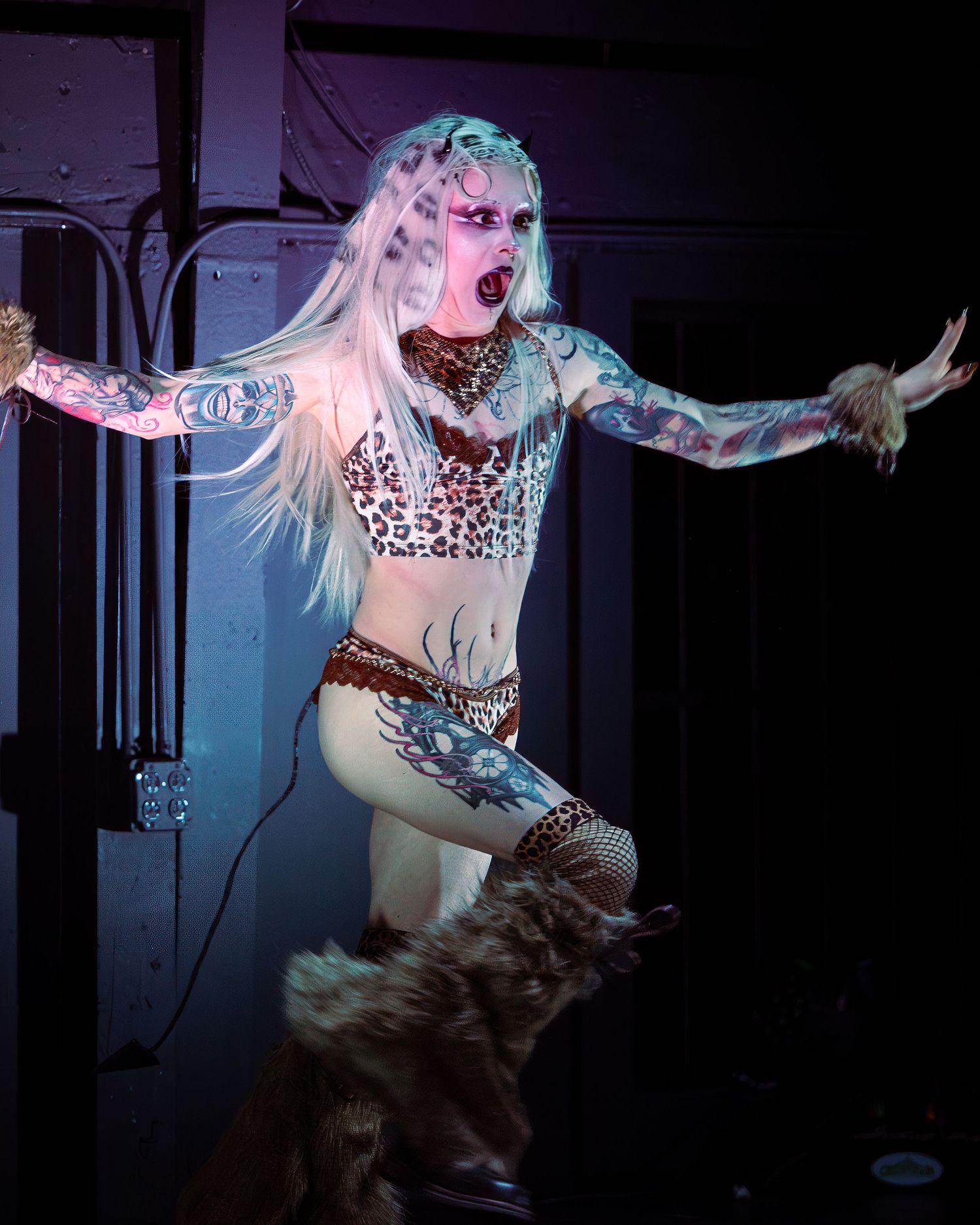Heavily tattooed blonde performer in leopard print outfit poses dramatically on stage under blue lighting.