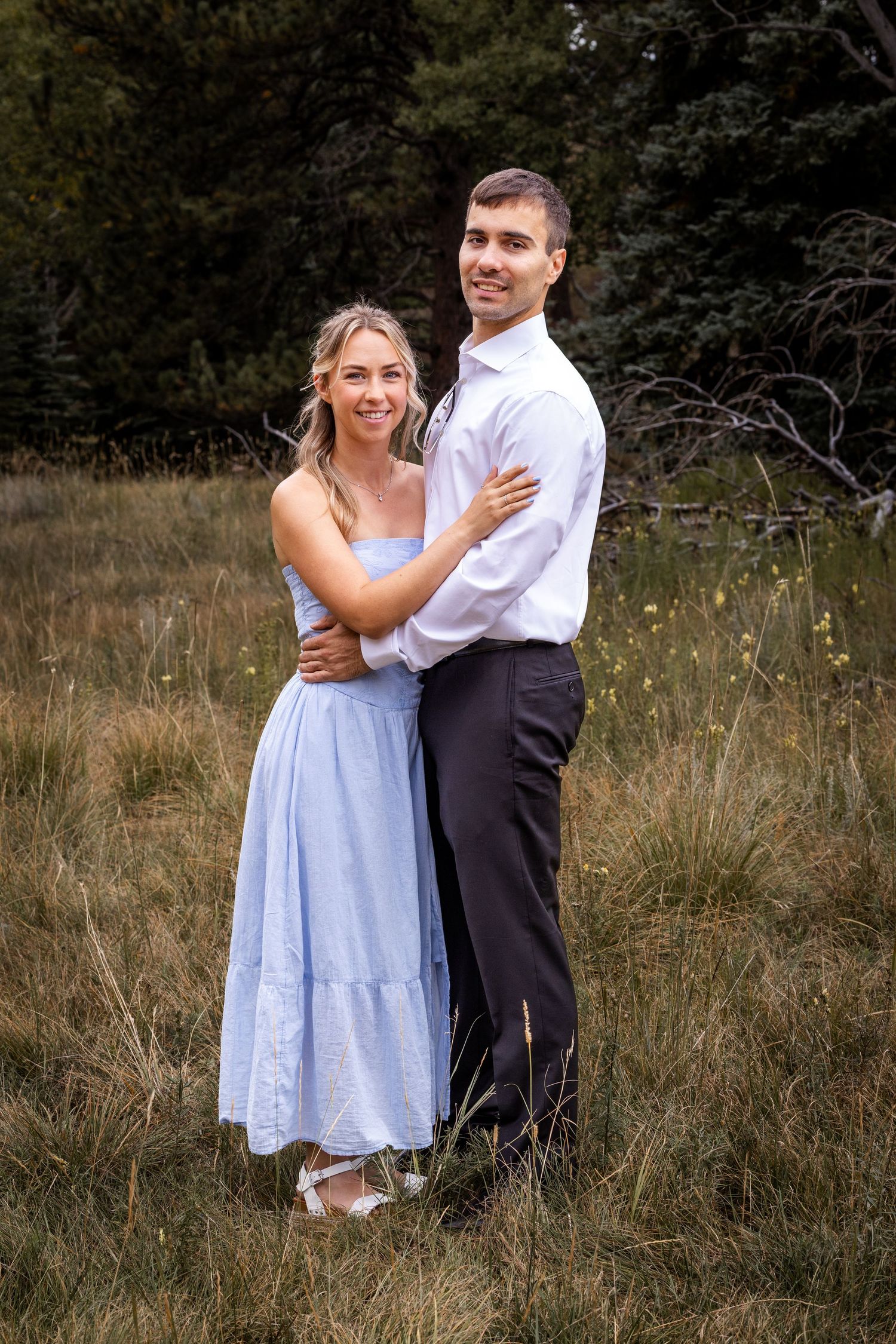 Romantic couple in formal attire embrace in a rustic outdoor meadow setting.