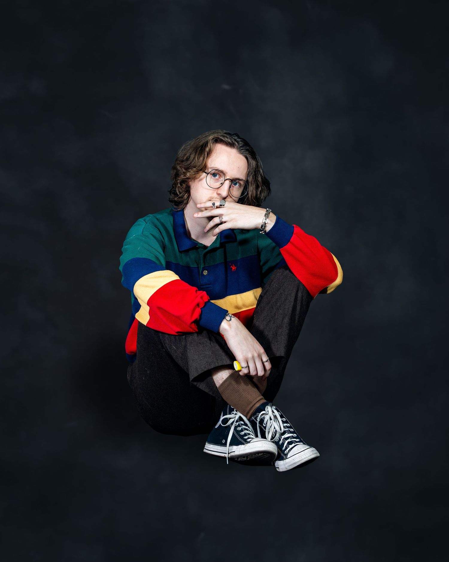 Young person in colorful color-block sweater and Converse sneakers sitting on floor, posing thoughtfully against dark background.