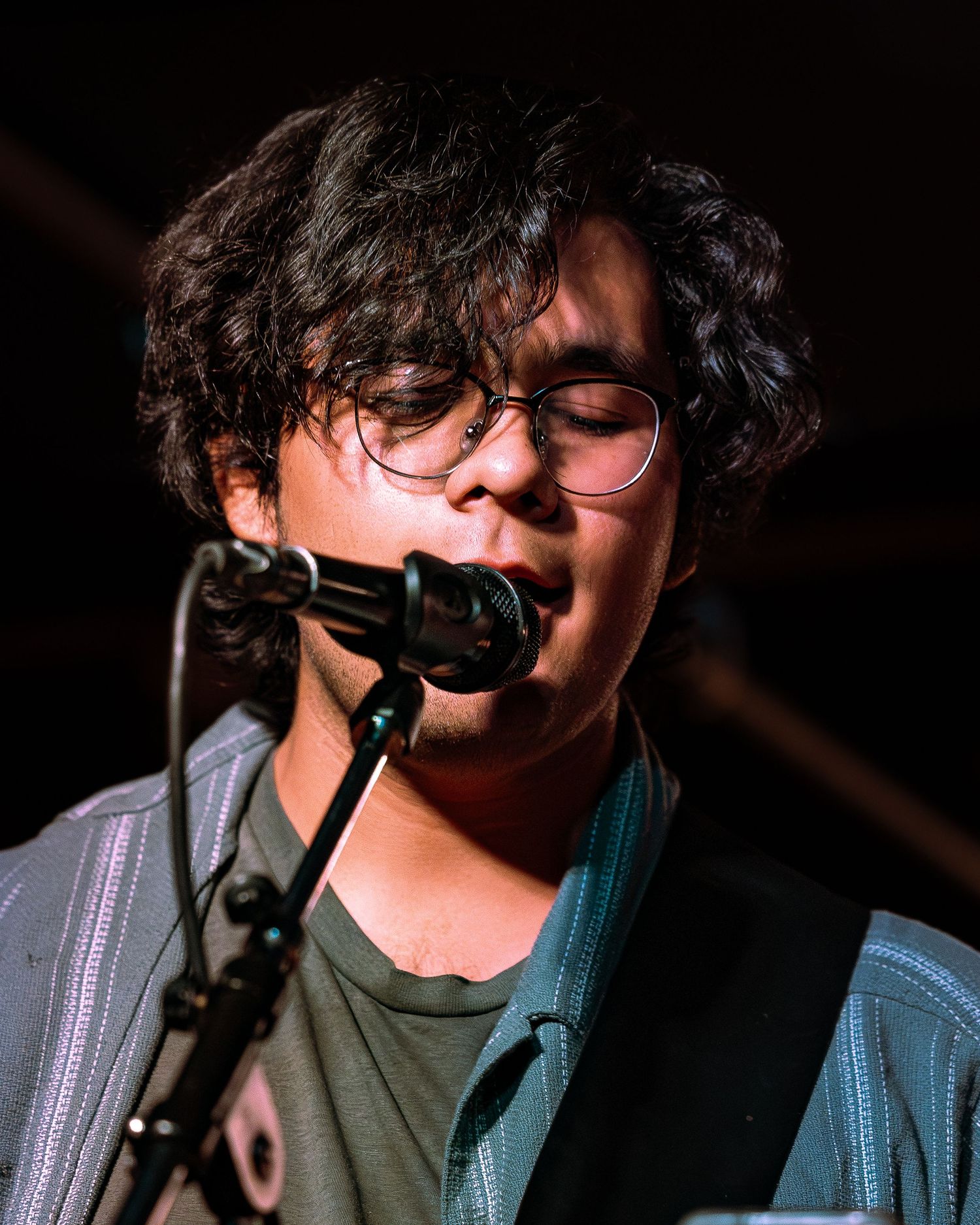 Young musician with curly hair and glasses singing into microphone while playing guitar on stage.
