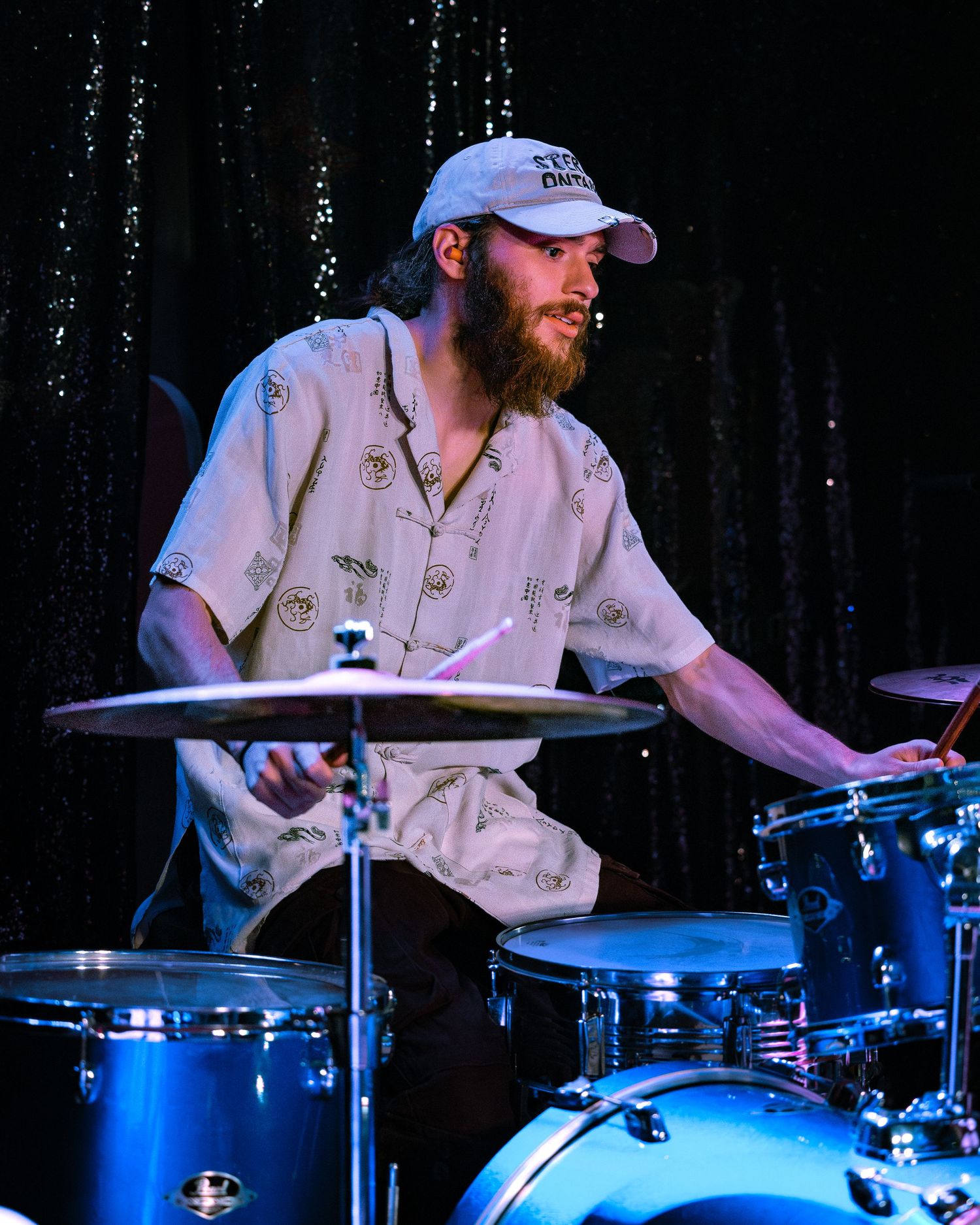 Bearded male drummer performing on stage with blue drum kit, wearing white cap and patterned shirt.