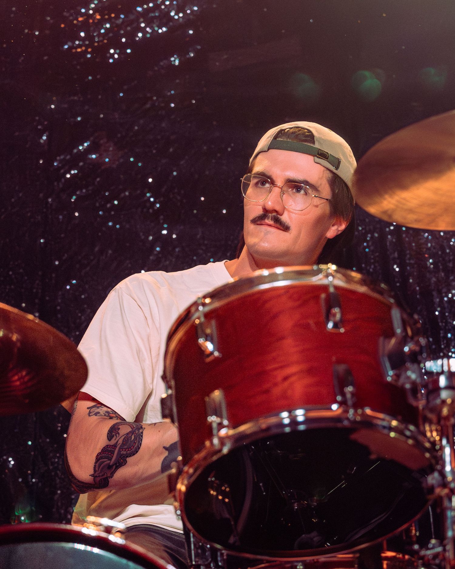 Drummer with mustache and backwards cap plays red drum kit under starry stage backdrop.