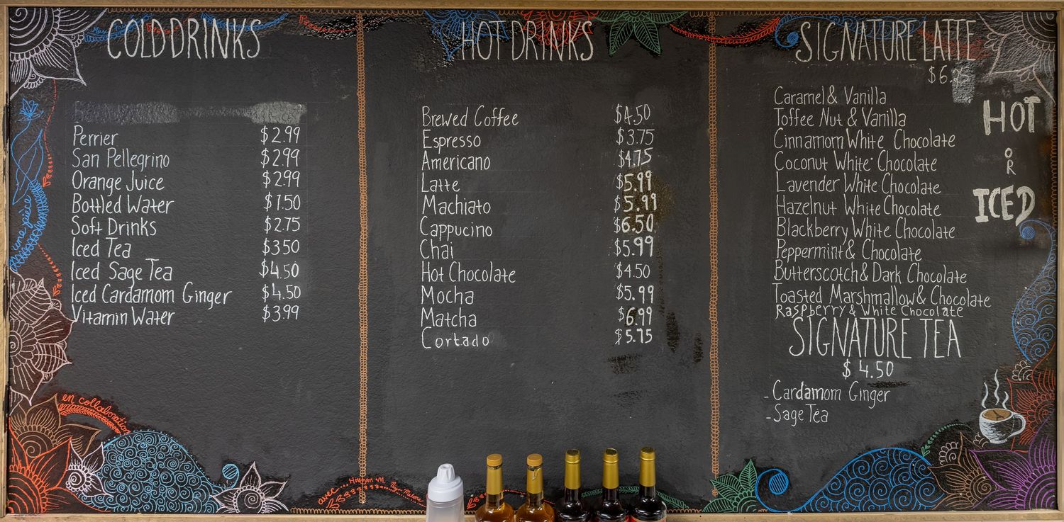 Chalkboard menu displaying cold drinks, hot drinks, and signature lattes with colorful hand-lettered text at a cafe.