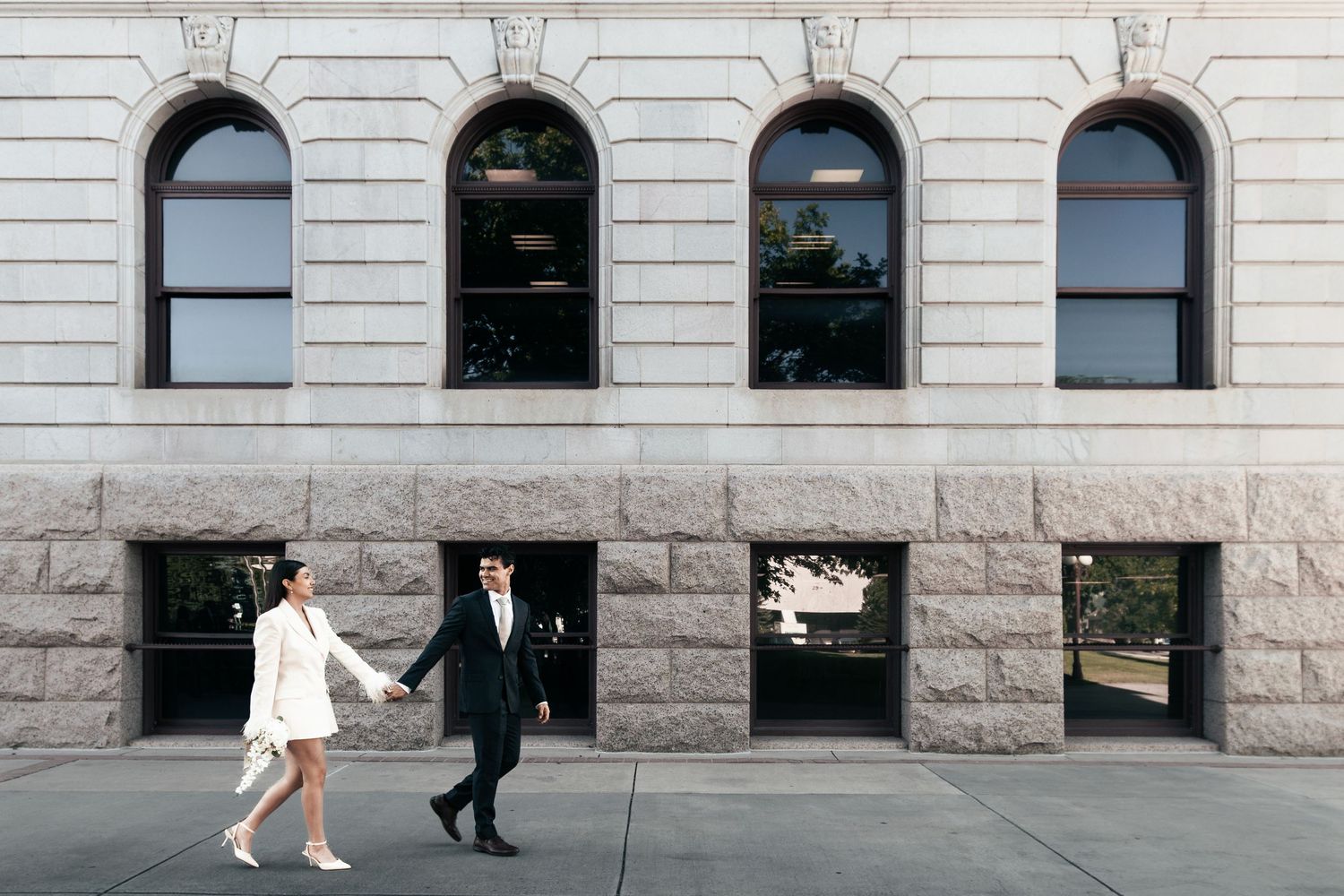 Couple in wedding attire holding hands walking past a grand stone building with arched windows.