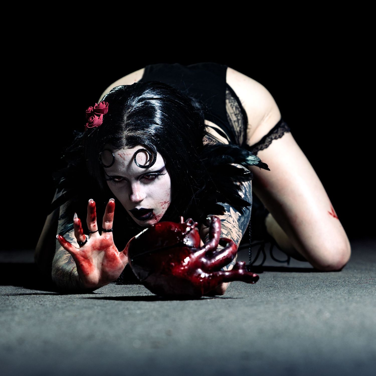 Gothic woman in dark makeup crawling on floor with bloody hands and heart prop in dramatic horror-themed photo shoot.