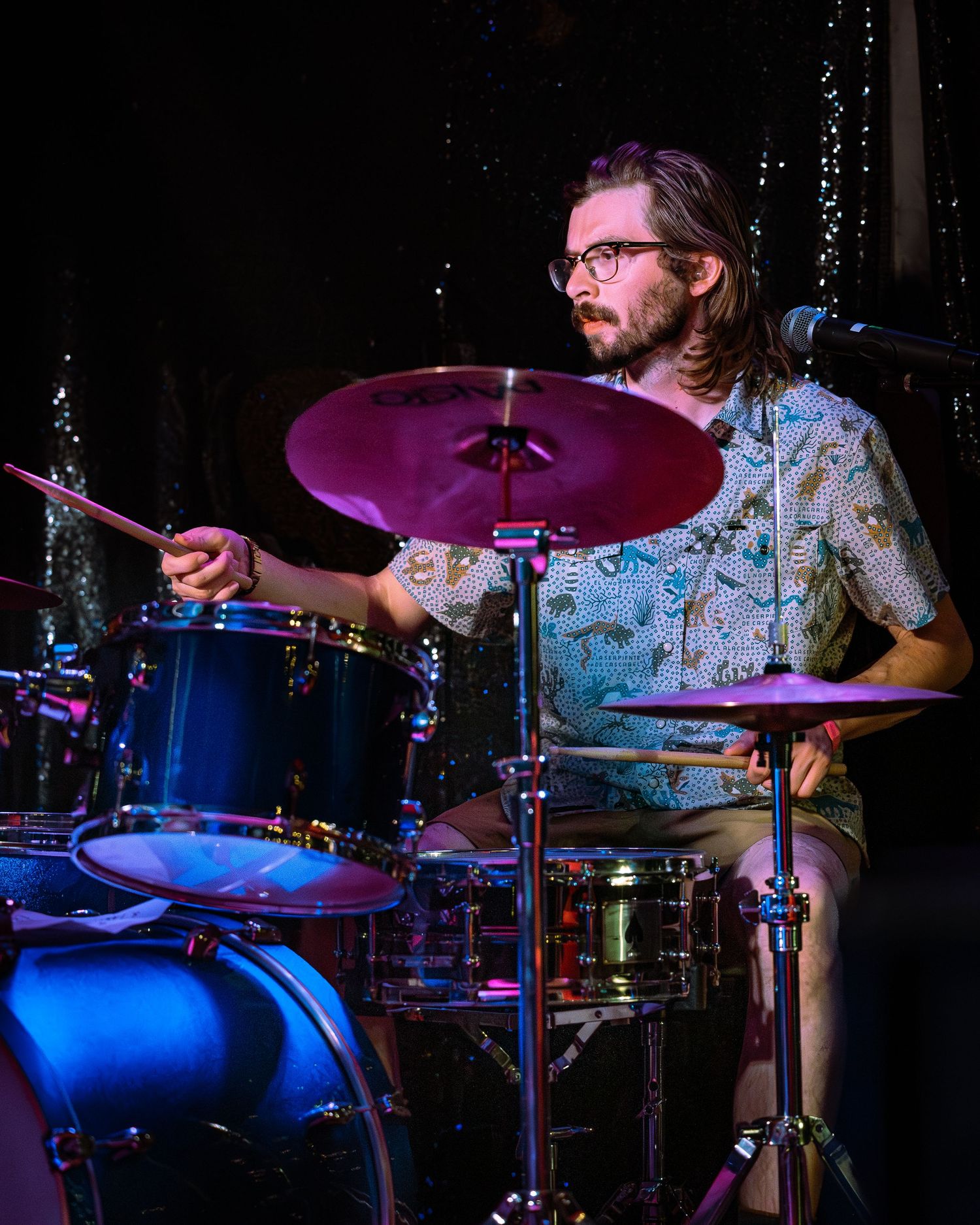 Bearded drummer in floral shirt plays drum kit under colorful stage lighting.