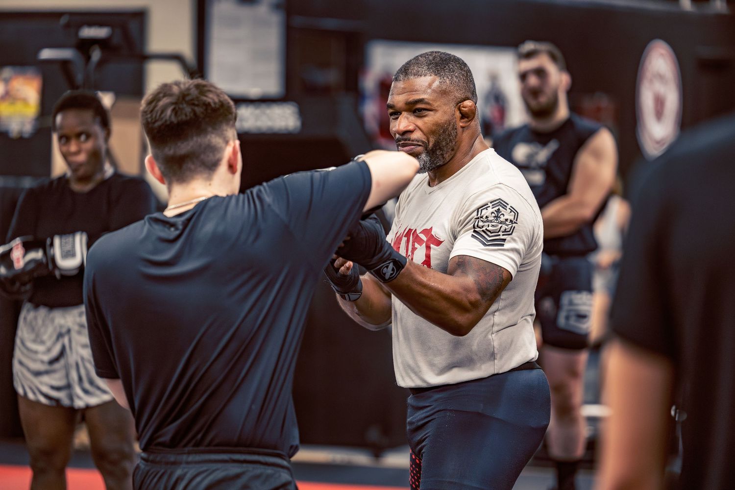 Martial arts practitioners engaging in combat training drills on red mats in a well-equipped gym.