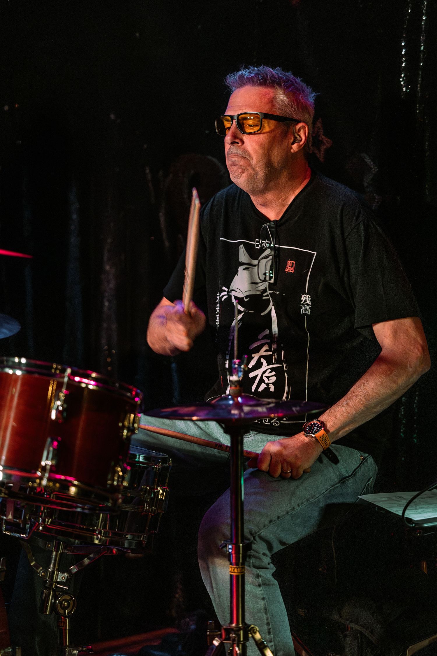 Drummer with orange sunglasses playing drums on a dark stage.
