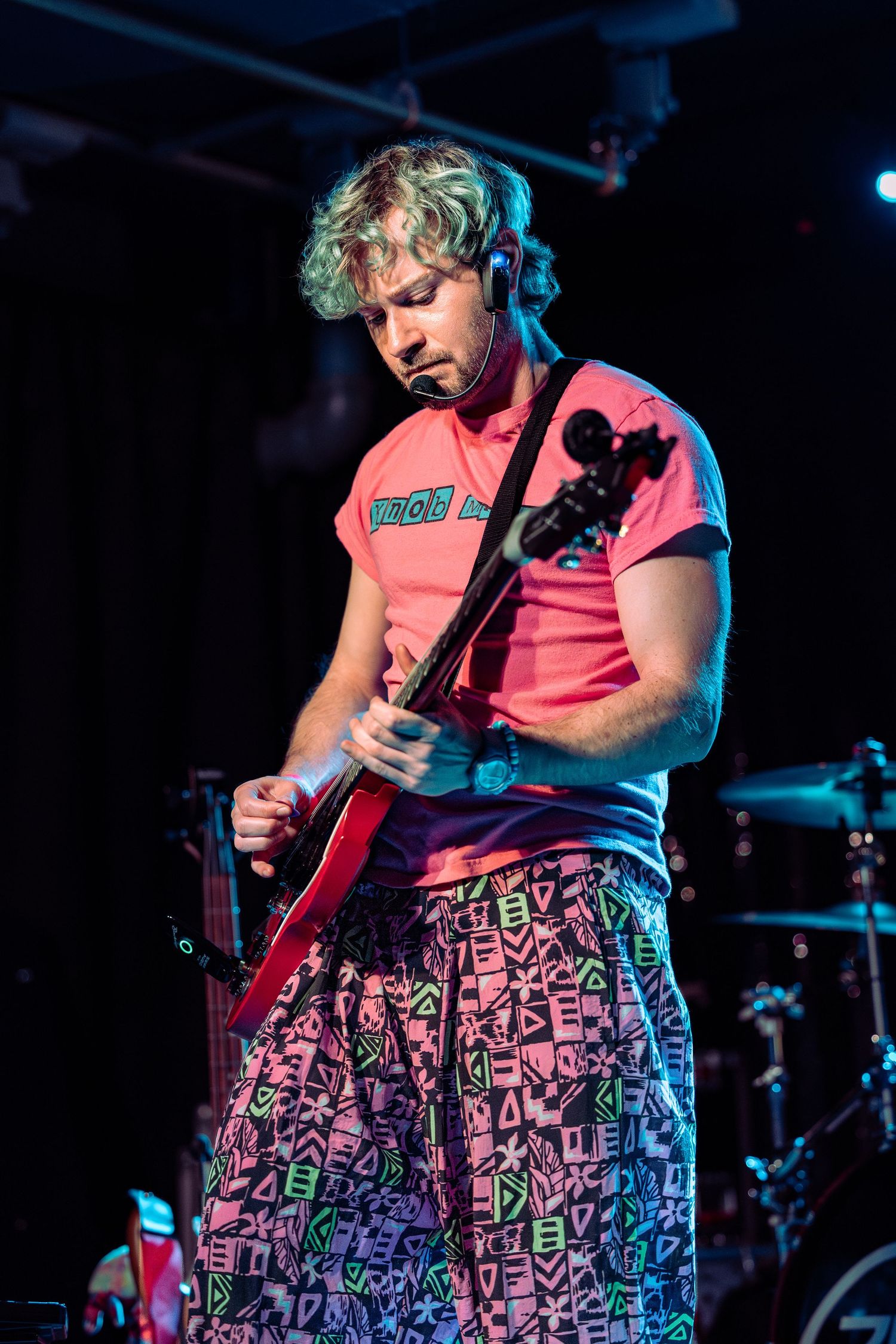 Blond guitarist in pink shirt and colorful patterned pants performs on stage under blue and pink lights.