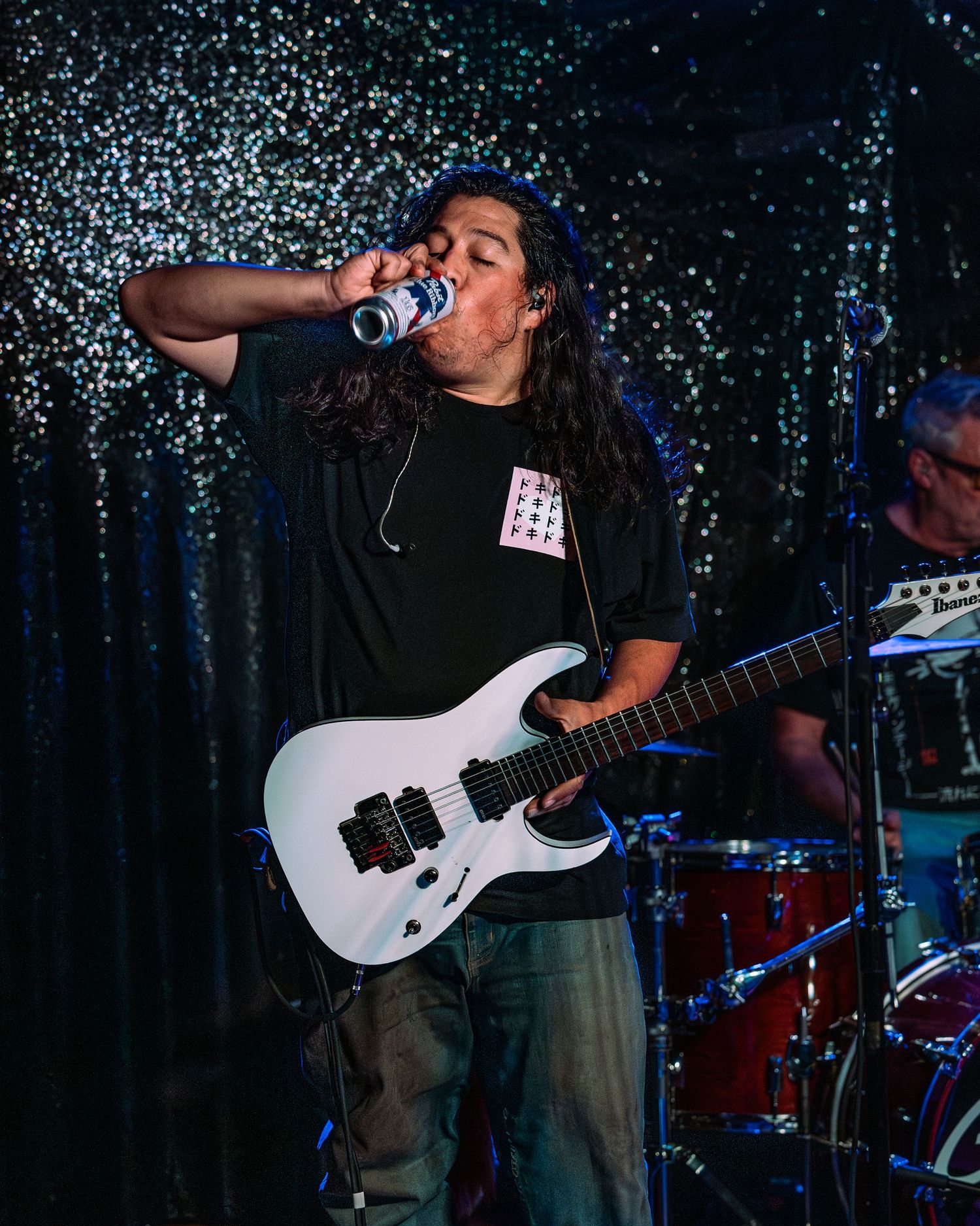 Long-haired guitarist drinking from can while holding white electric guitar on glittery stage.