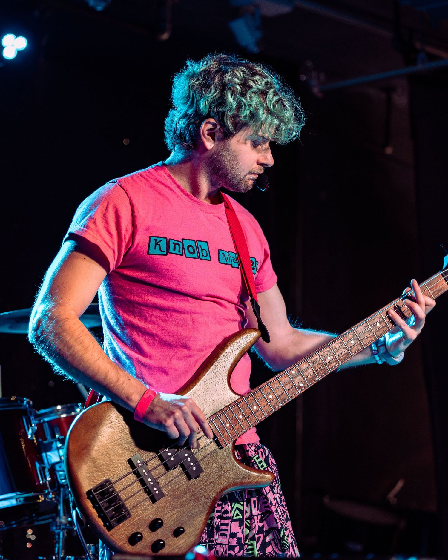 Curly-haired man in pink t-shirt playing brown bass guitar on dark stage with blue lighting.
