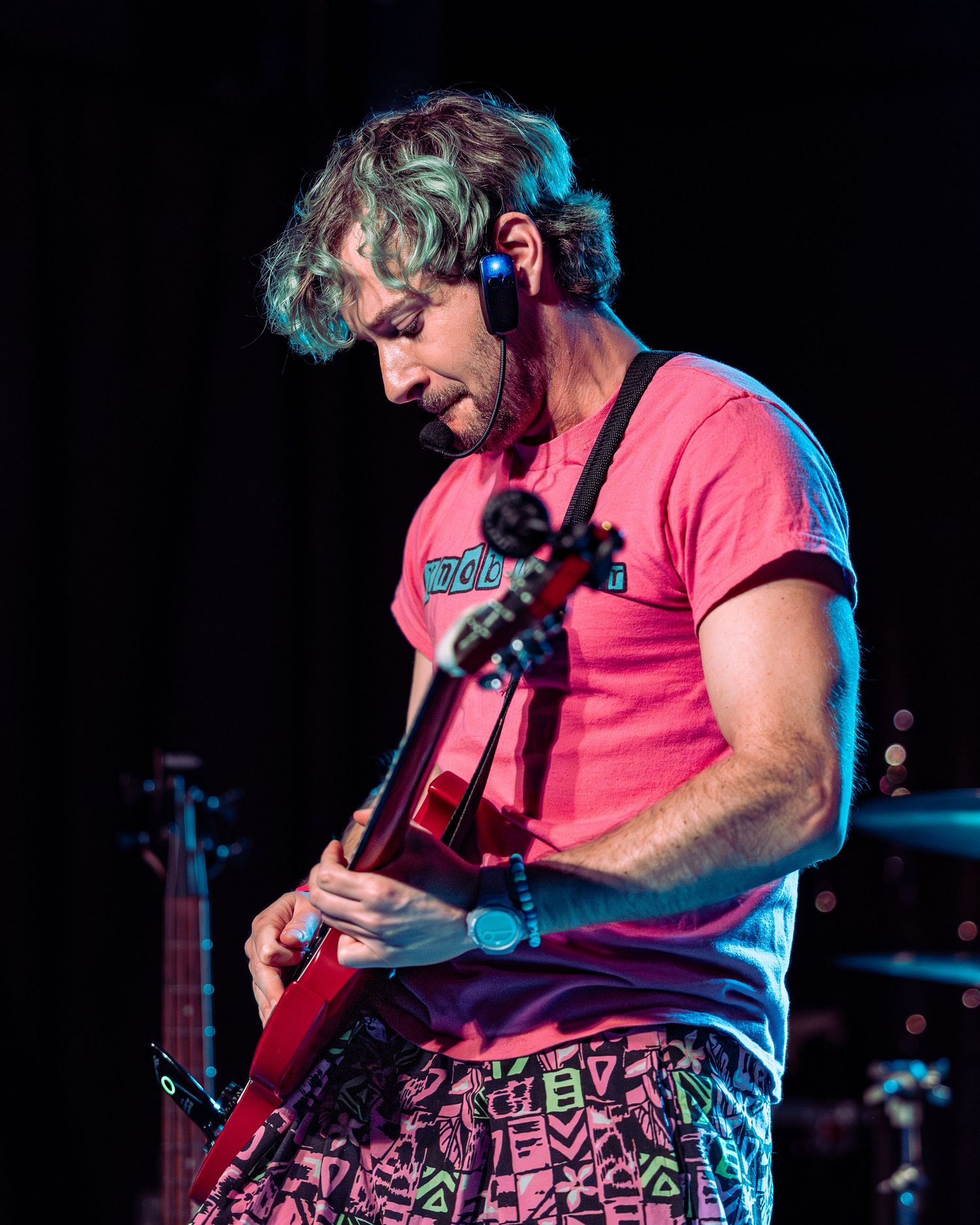 Musician in pink shirt plays red electric guitar on stage under blue and pink stage lighting.