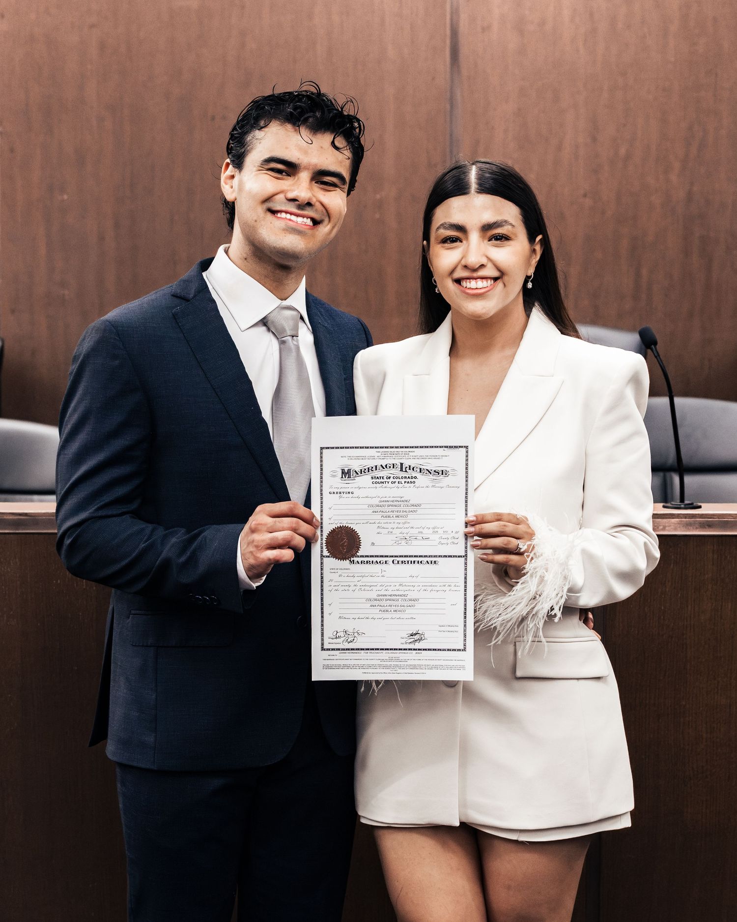Couple in formal attire smiling while holding a marriage license certificate in a courthouse.