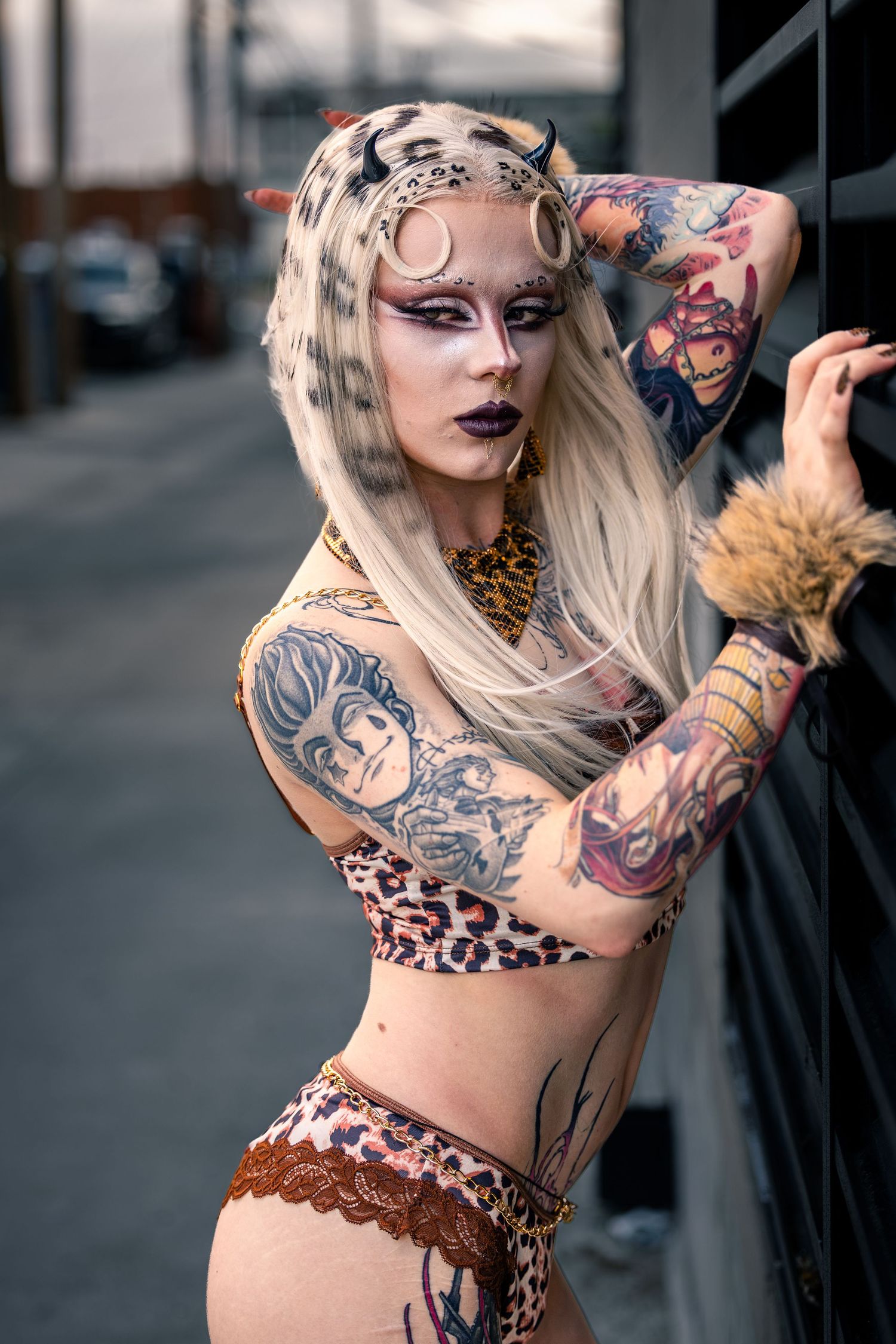 Tattooed woman in leopard print bikini with face paint and horns poses against urban wall.