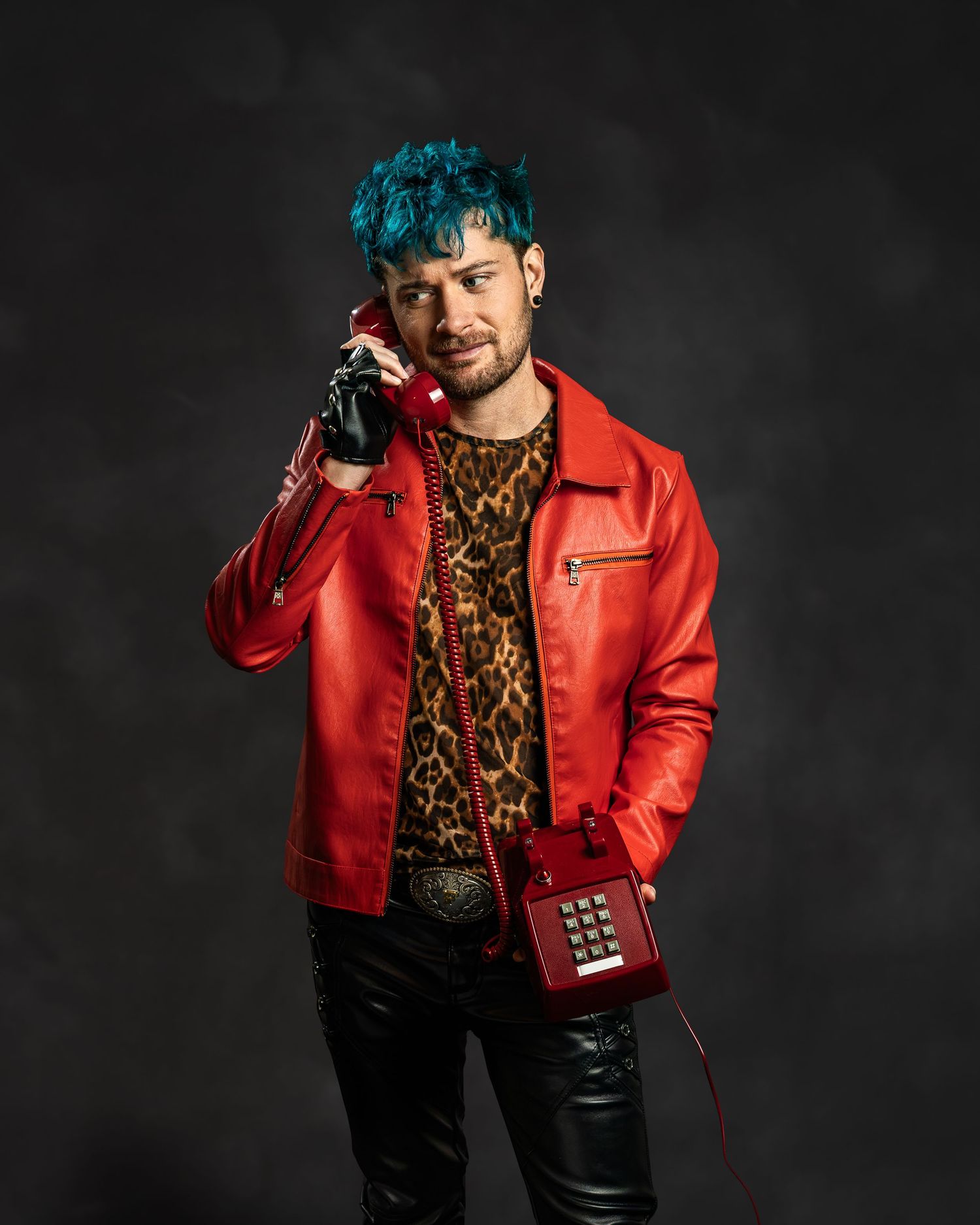 Man with blue hair in red leather jacket holding vintage red phone, wearing leopard print shirt and black pants.
