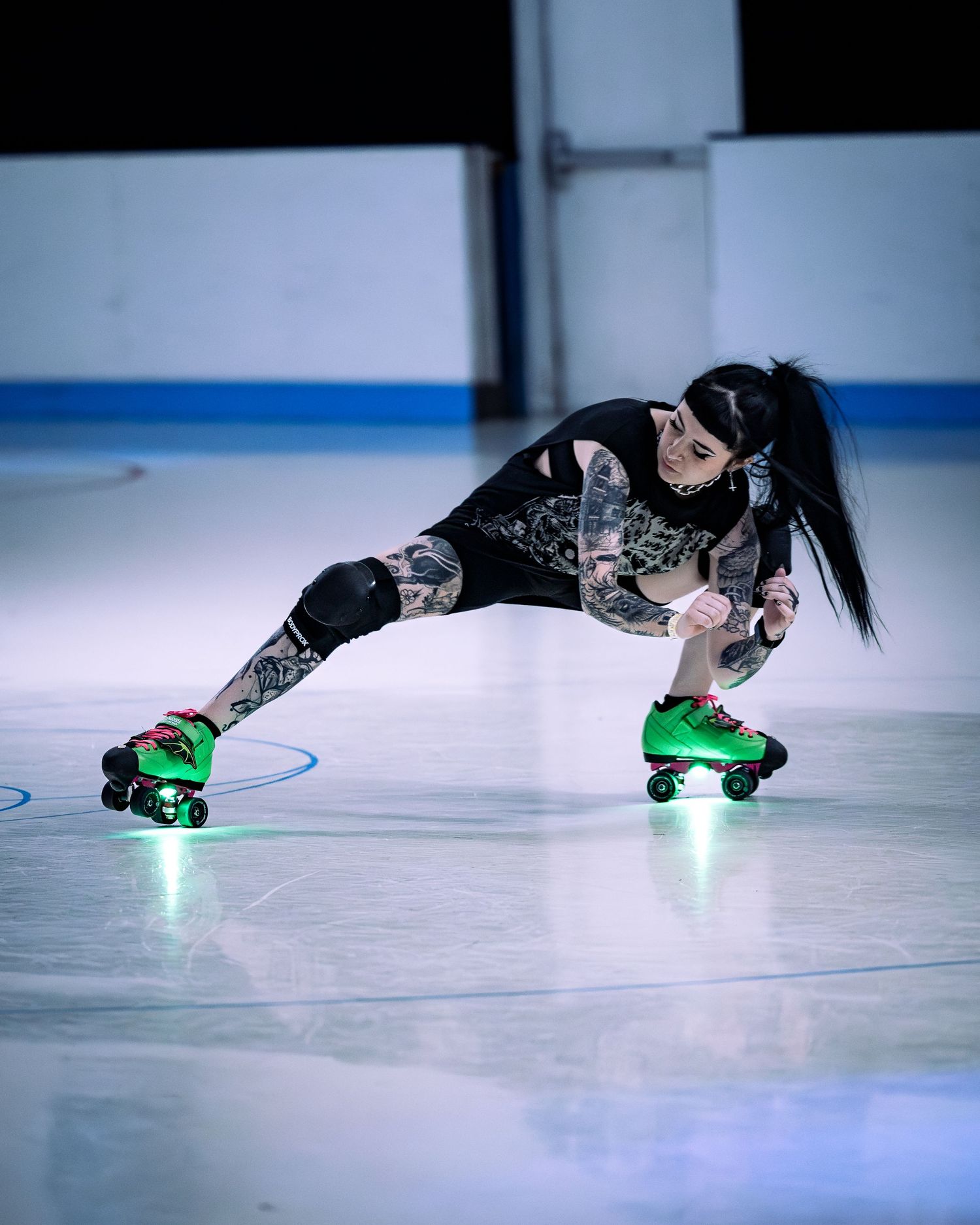 Tattooed roller derby skater in green skates crouches low on an ice rink in black athletic wear.