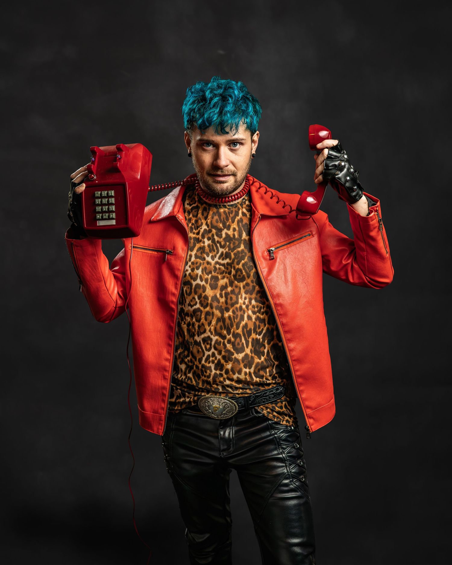 Man with blue hair in red leather jacket and leopard print shirt holds vintage red phone against dark background.