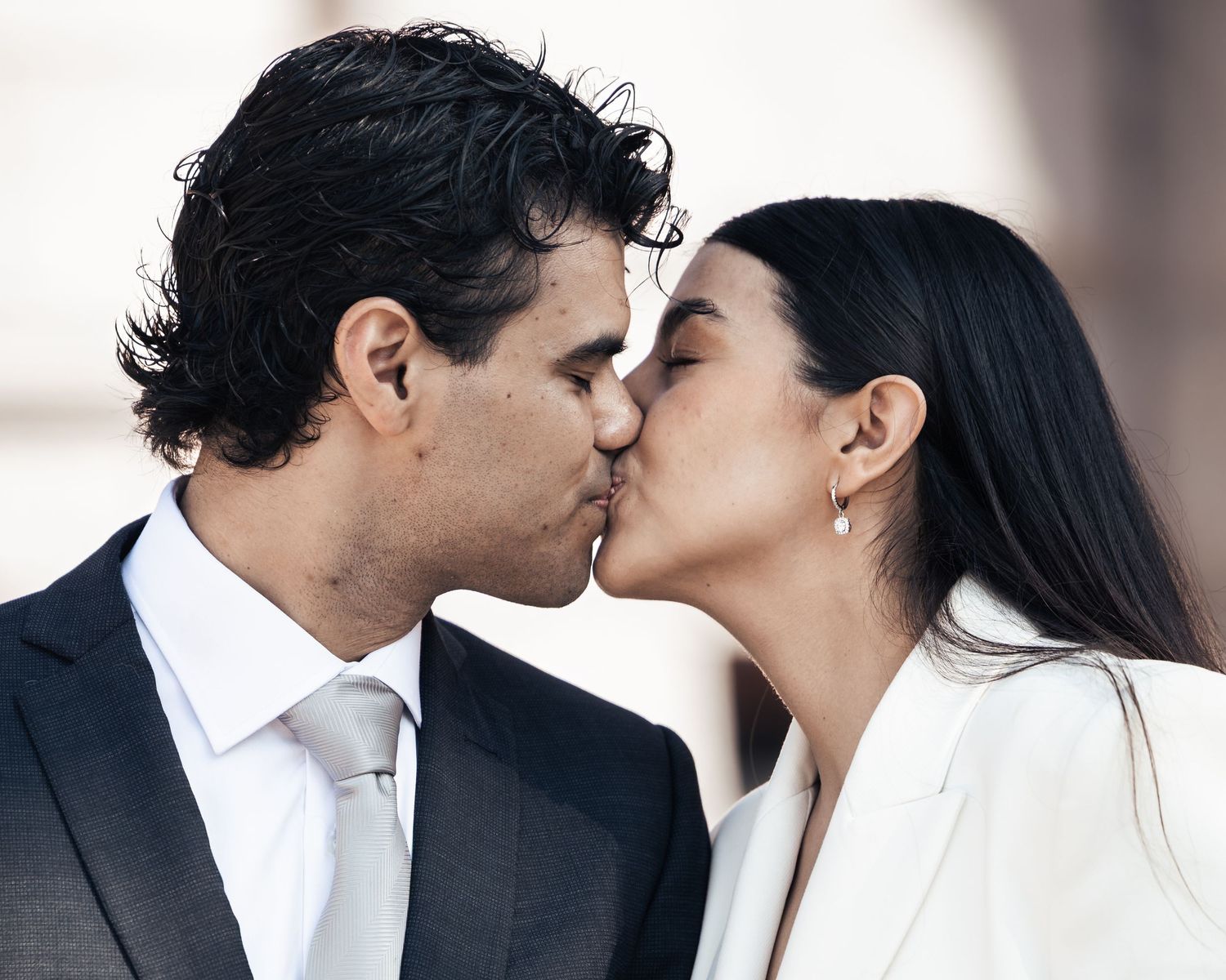 A couple shares a romantic kiss, the man in a dark suit with white tie and the woman in a white blazer.