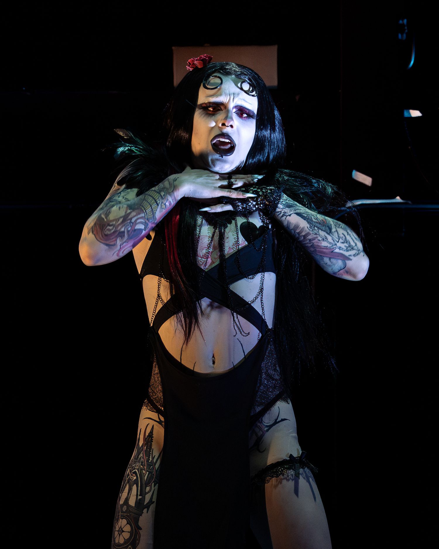 Gothic performer with heavy makeup and tattoos poses dramatically in black outfit on dark stage.