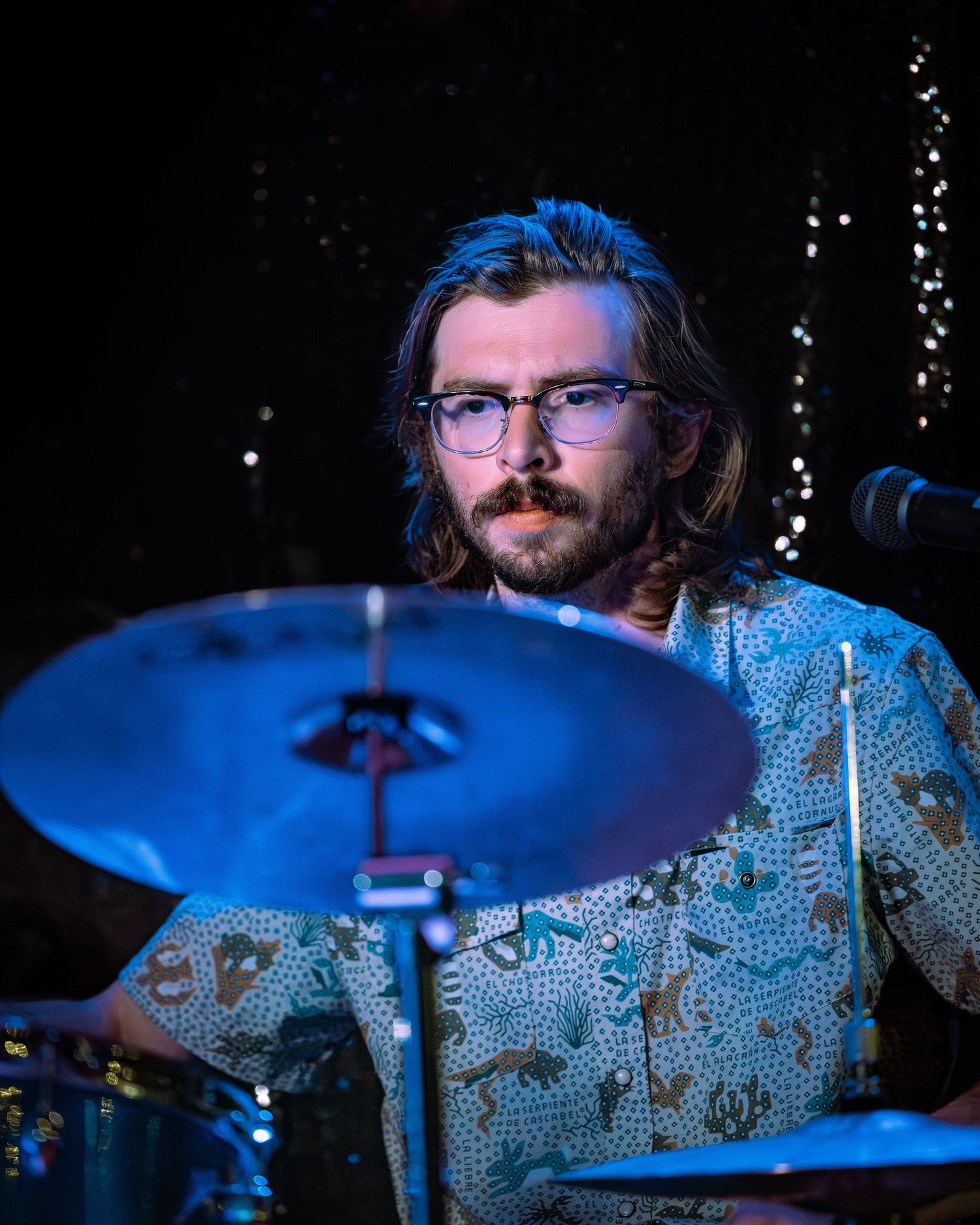 Bearded musician with glasses and shoulder-length hair playing cymbals on a blue-lit stage.