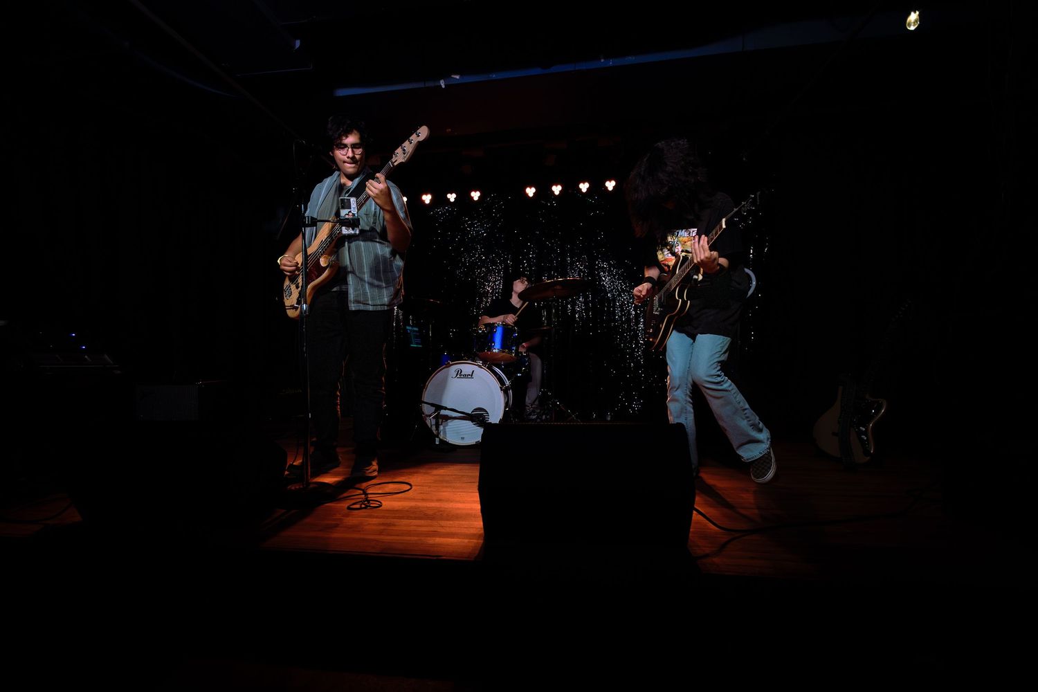 Live band performing on a dimly lit stage with guitarist, drummer, and vocalist under warm spotlights.