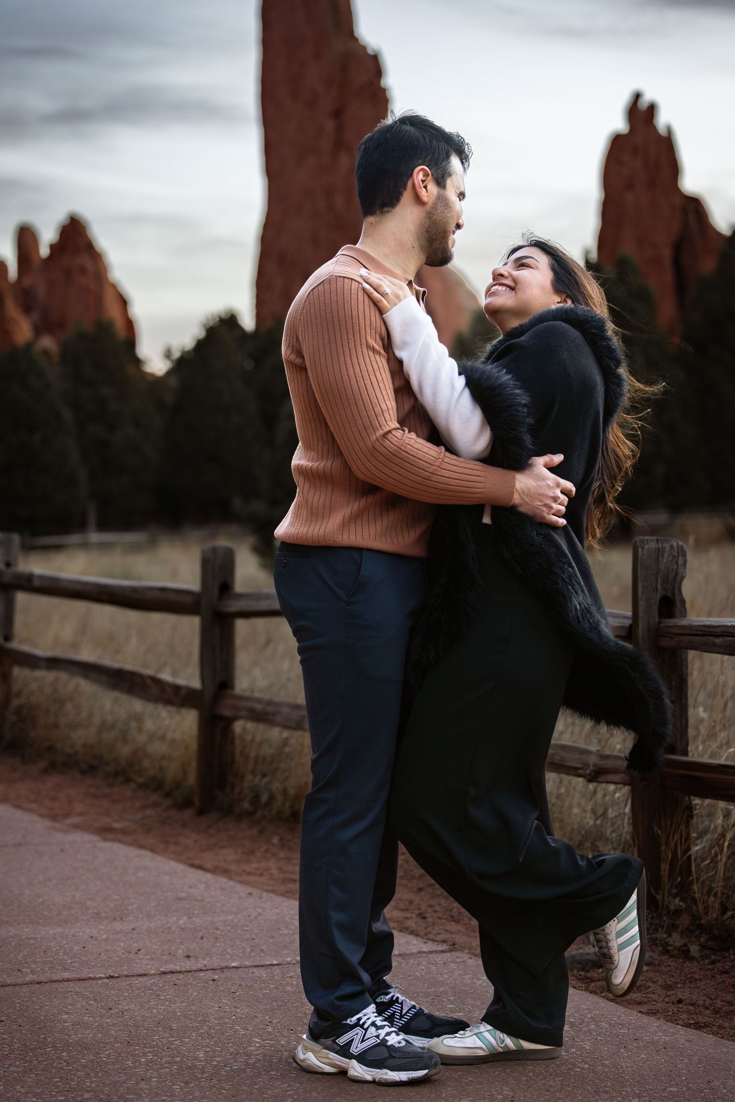 A couple embraces near a wooden fence during an outdoor photo session at sunset.